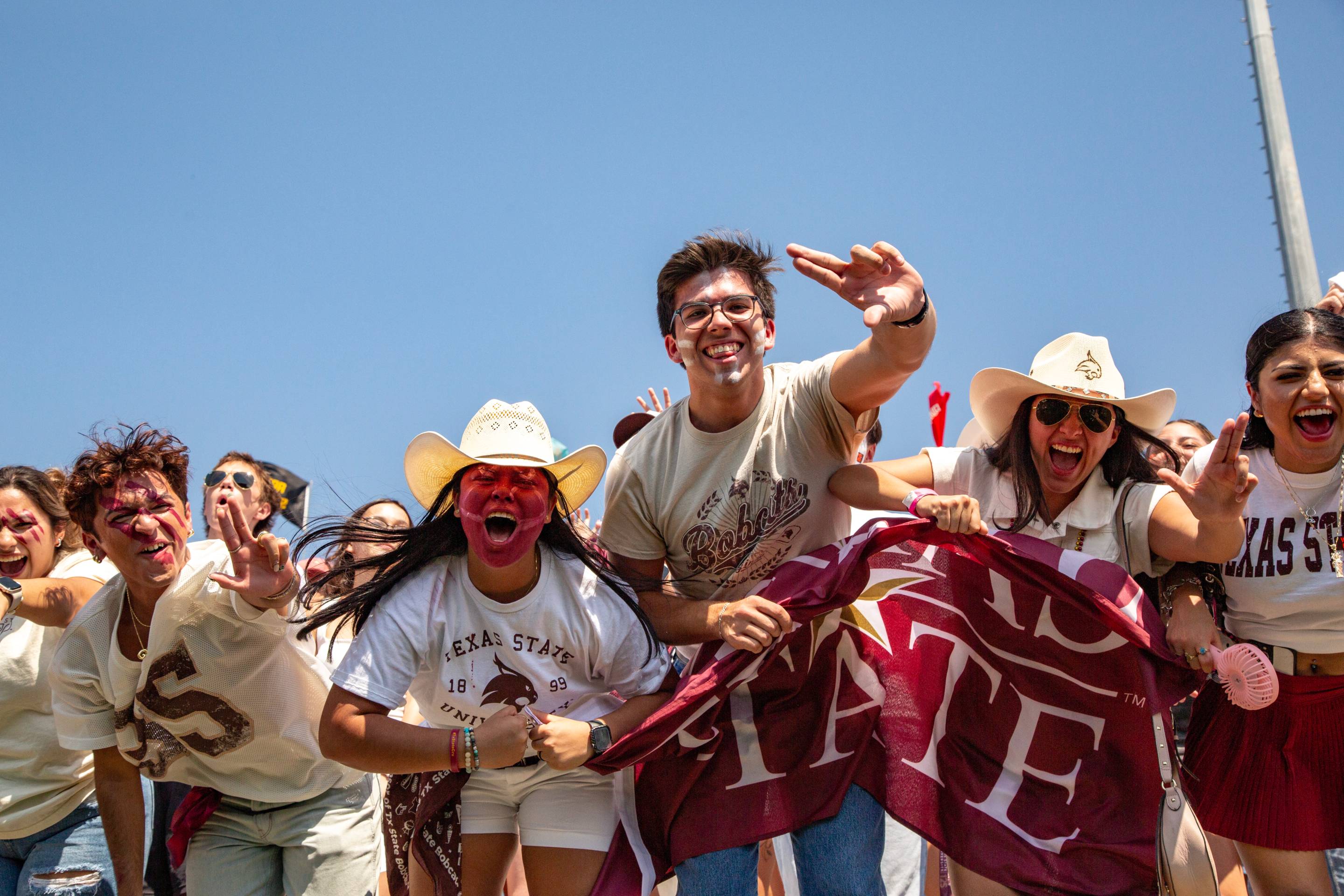 A group of students in TXST shirts, some with face paint, cheer and wave a TXST flag at a football game.