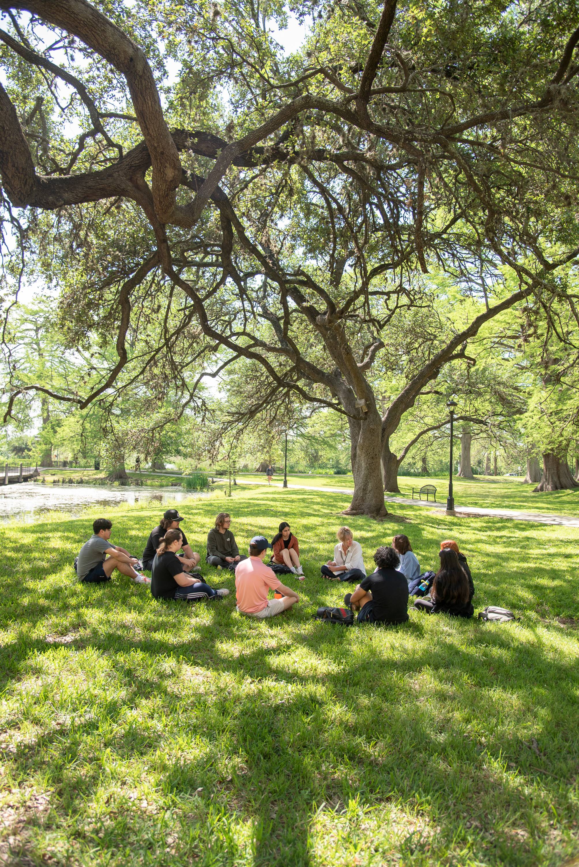 A small group of students participates in class outdoors by a pond and large tree.