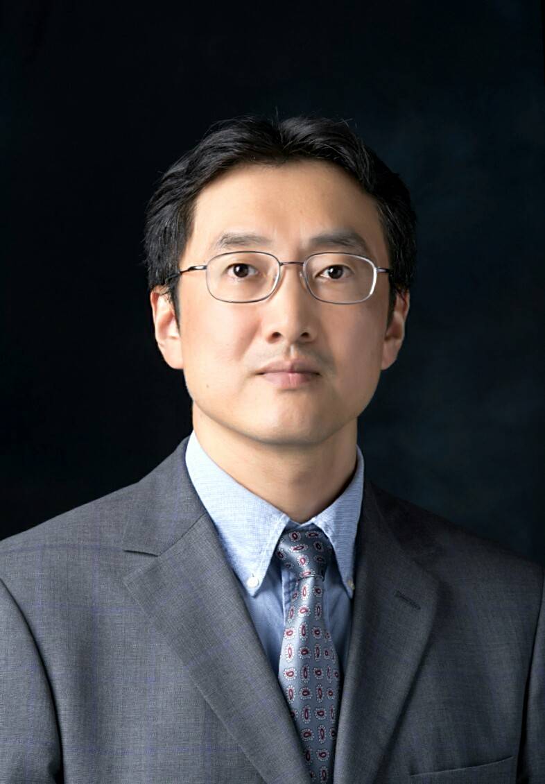 A professional headshot features a faculty with dark hair and glasses, wearing a grey suit, a light blue collared shirt, and a patterned tie, looking directly at the camera against a dark brown background.