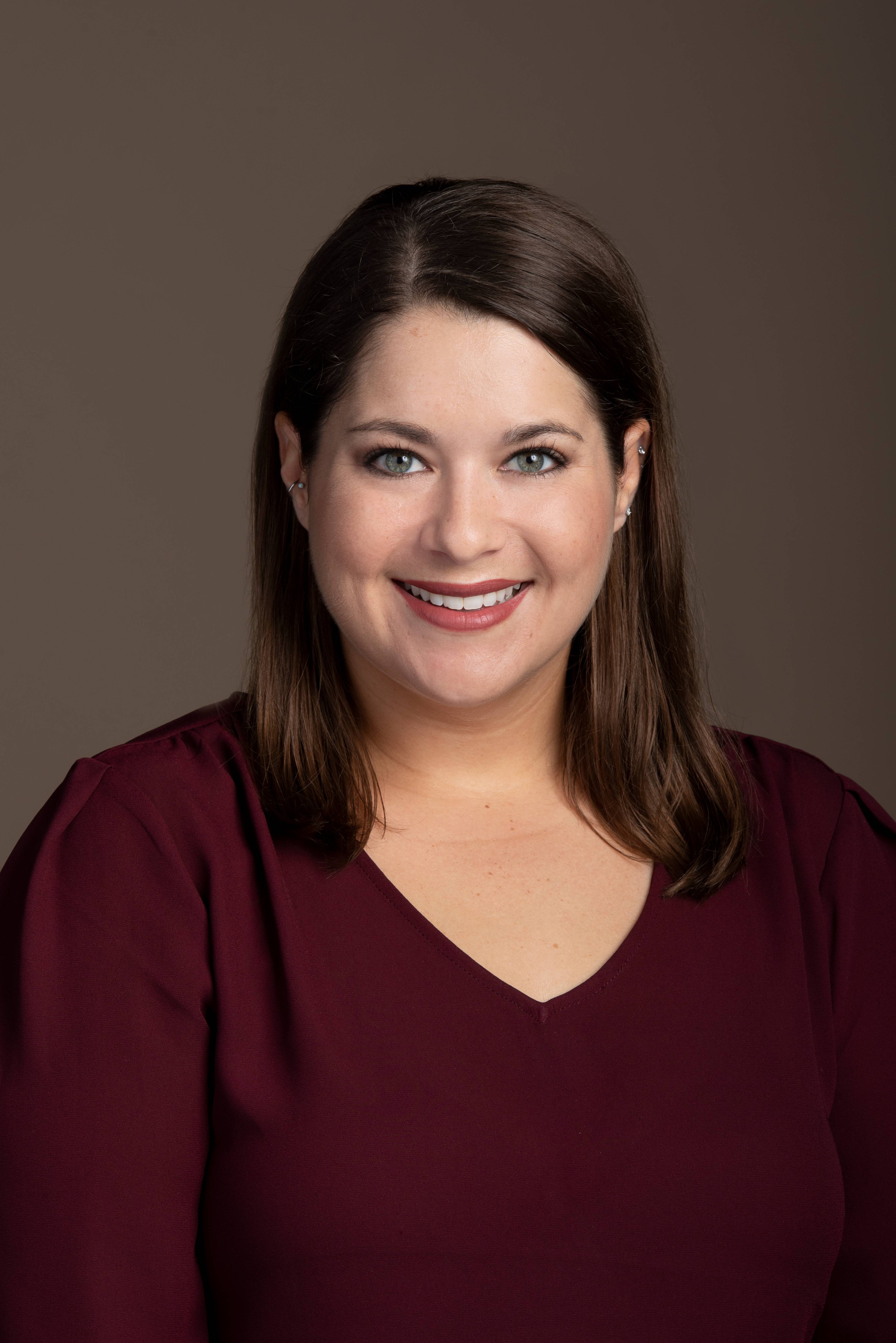 A headshot of a smiling faculty with shoulder-length brown hair and light eyes, wearing a burgundy V-neck top, against a muted brown background.