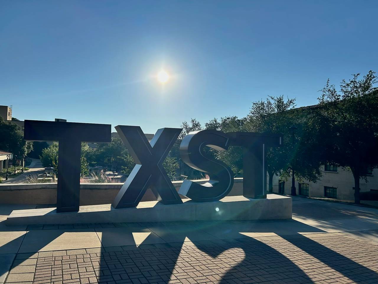 Photo of the TXST sign near the Quad with the sun shining in the background.