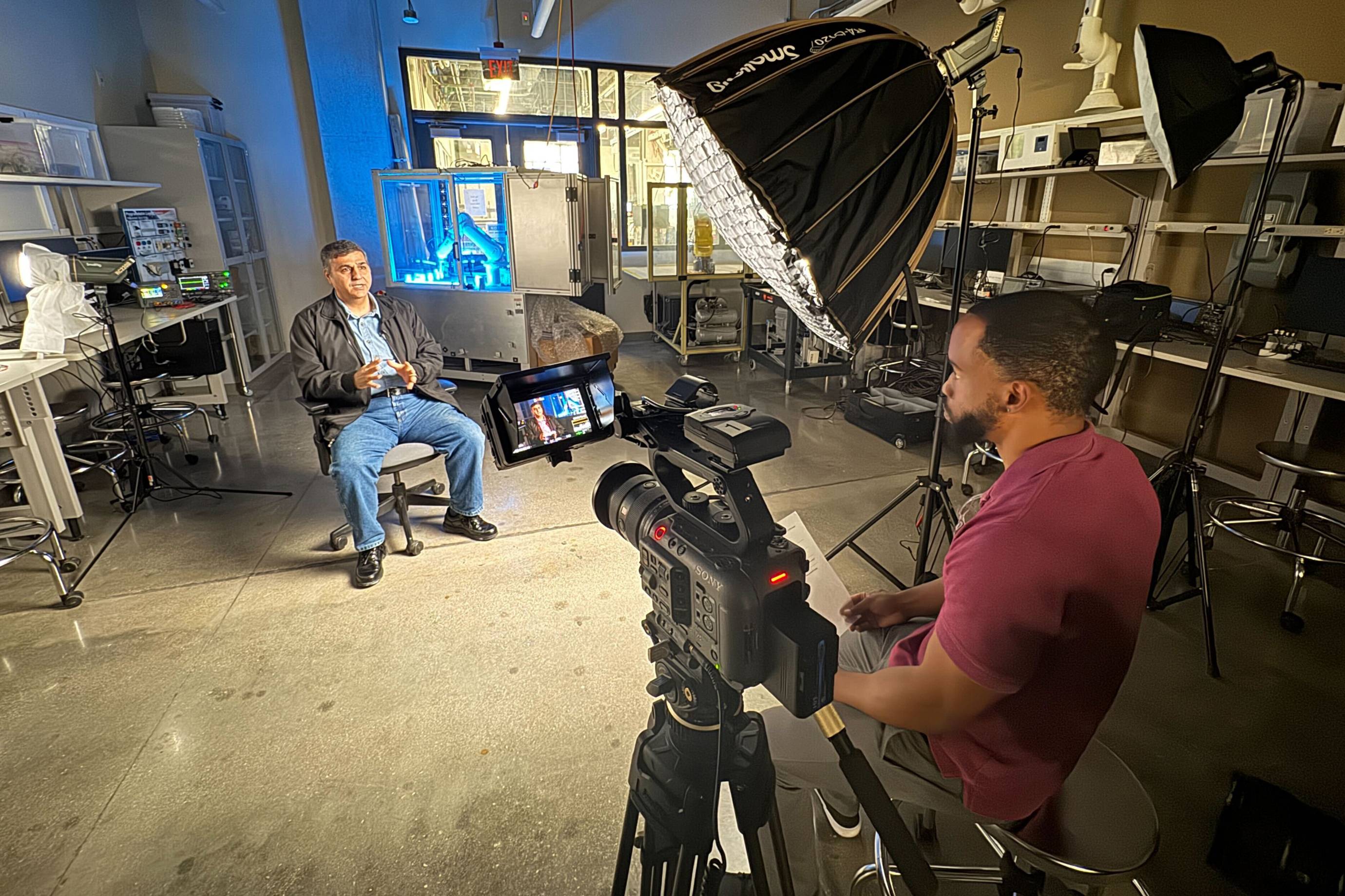 man in black jacket sitting in lab setting while getting interviewed on camera.