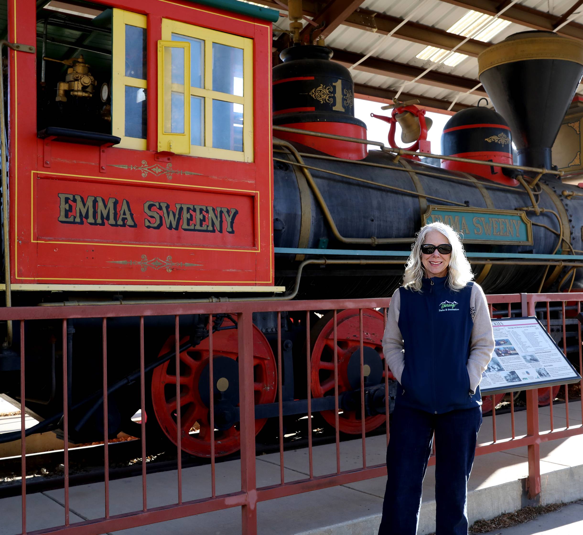 woman in blue vest stands next to red locomotive that reads "emma sweeny"
