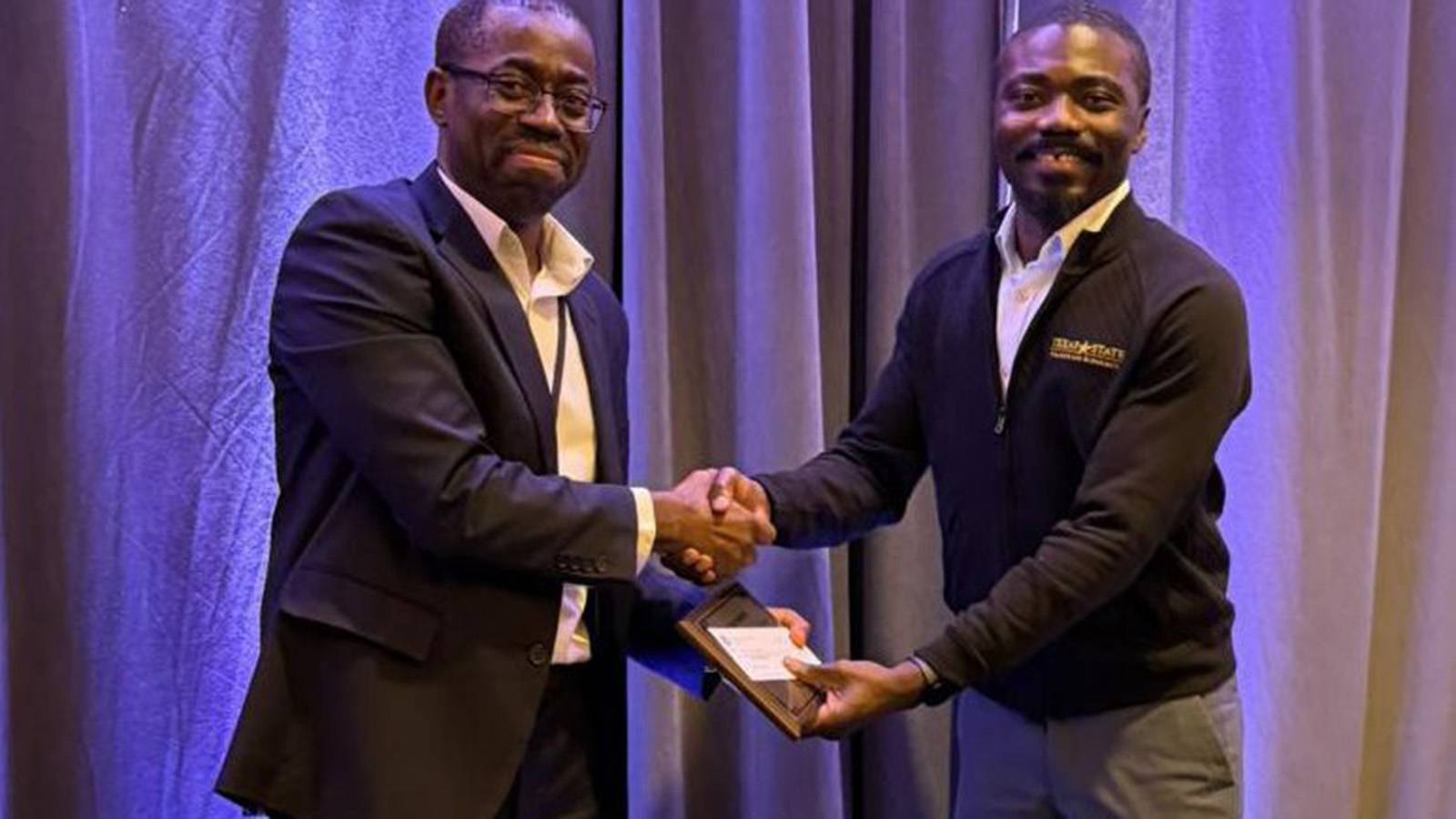 two men dressed in business attire, shaking hands while one hands the other an award