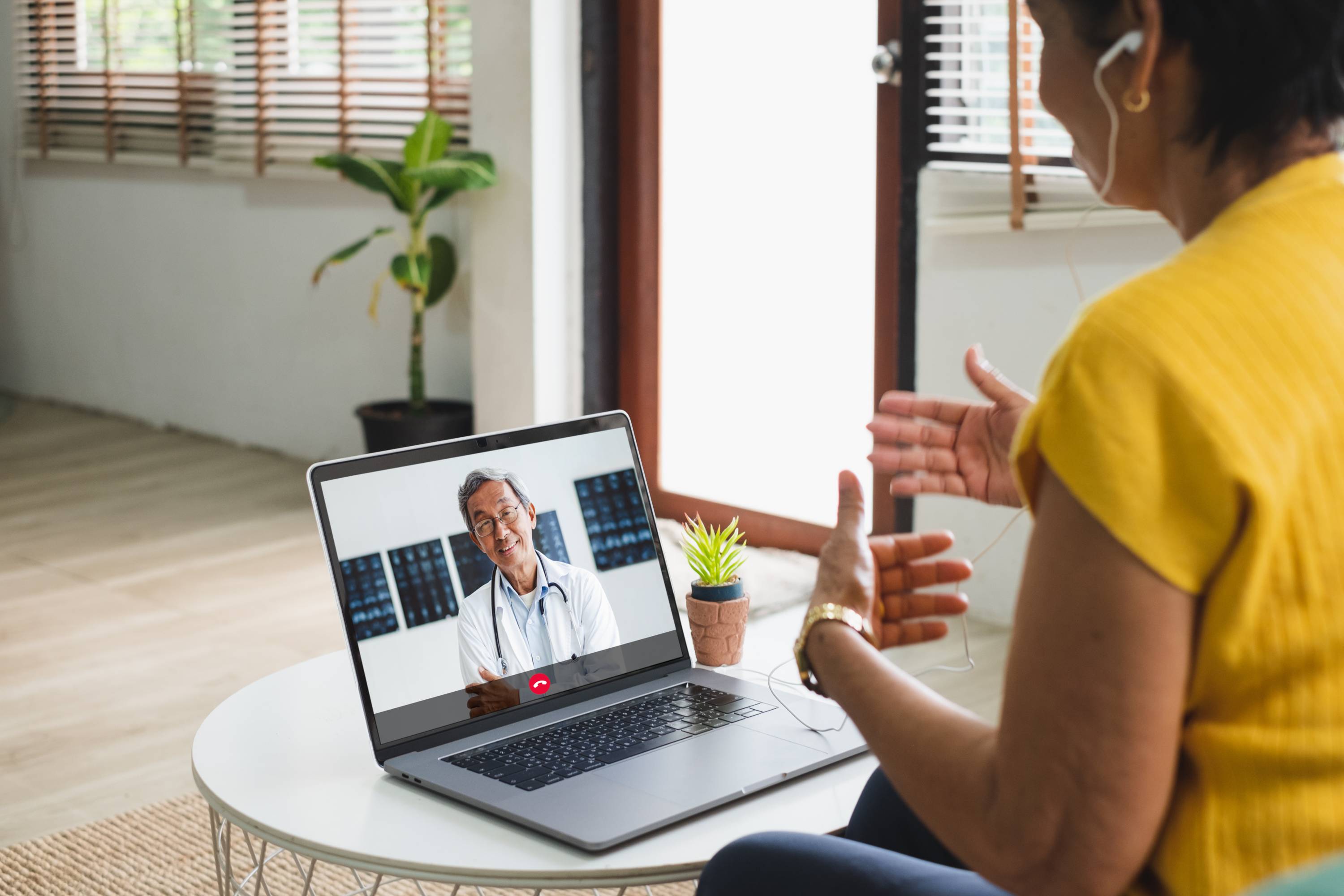 A woman an yellow is having a telehealth appointment with a doctor on her laptop.