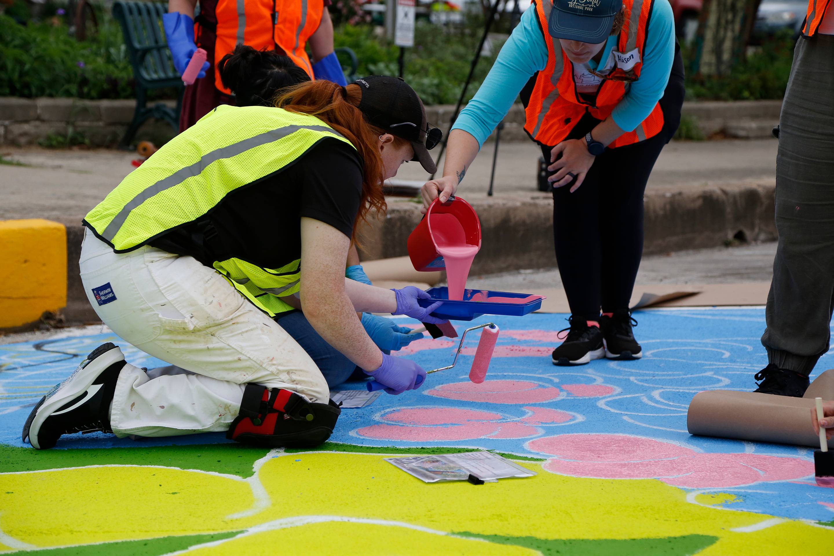 woman is pouring pink paint in another woman's paint palette. they are both wearing safety vests