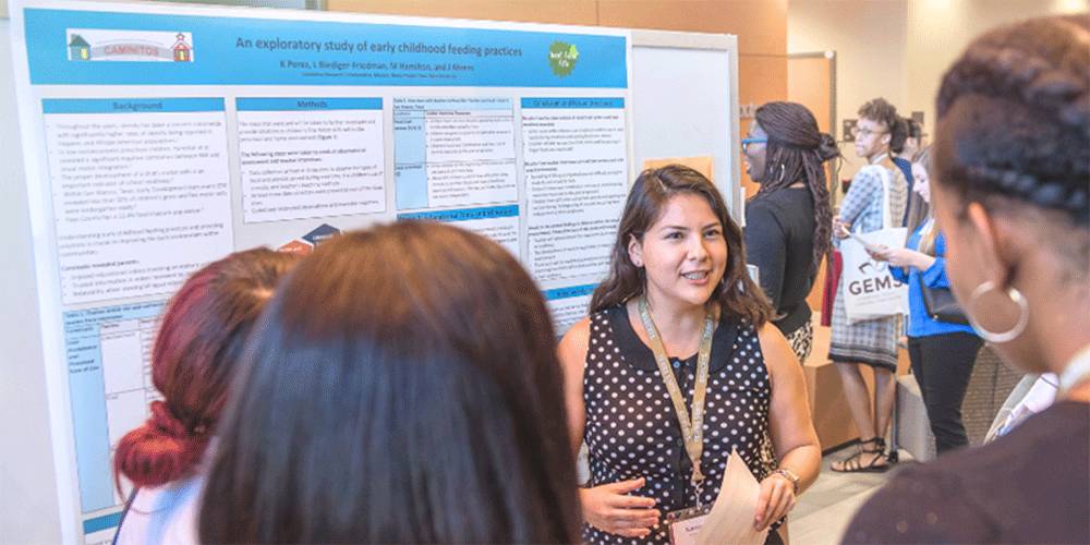Presenter passionately explains her research in front of a poster to a crowd of attendees at a conference
