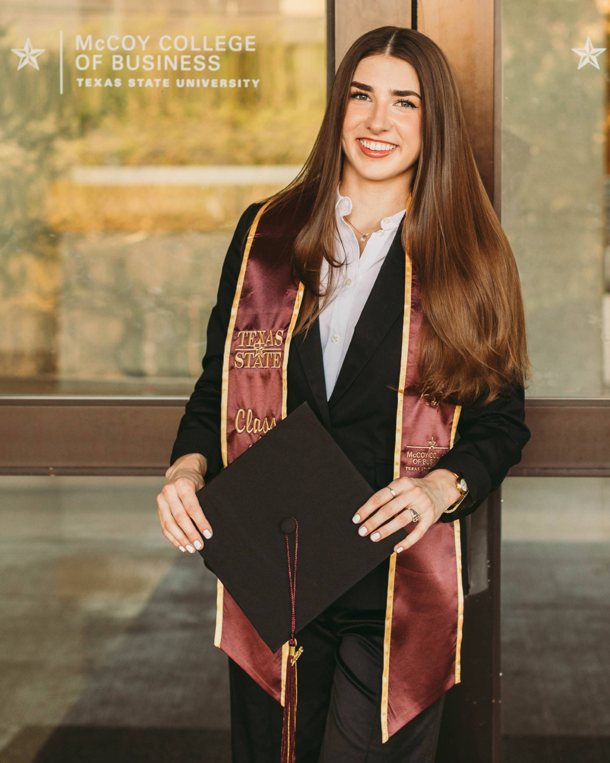 Young woman in graduation regalia posing outside doors of McCoy Hall