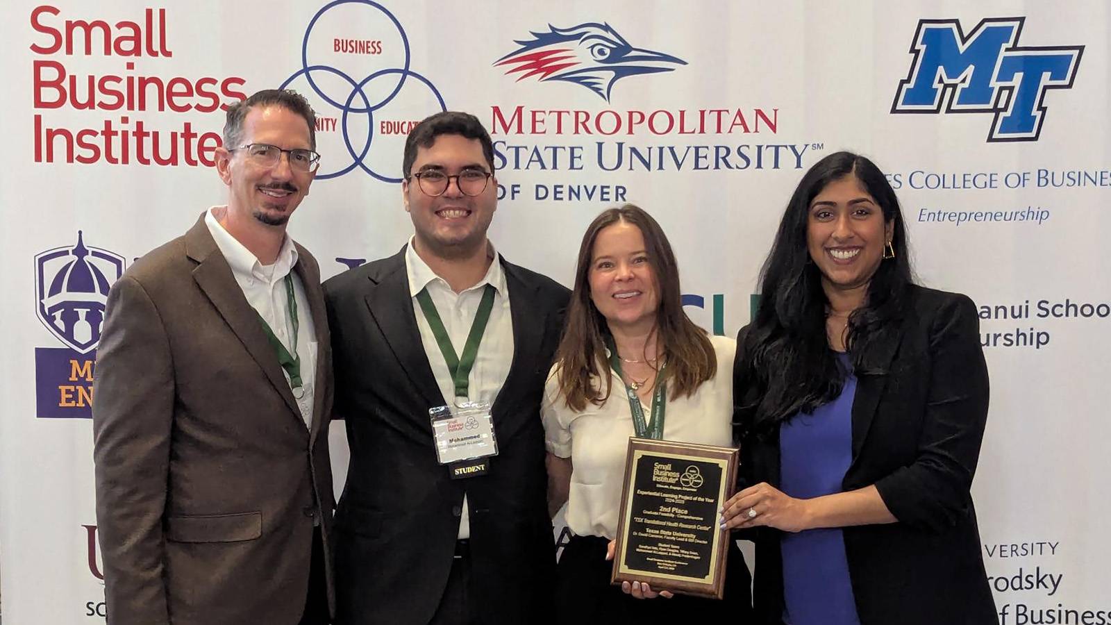four professionally dressed graduate students pose for photo while holding award plaque 