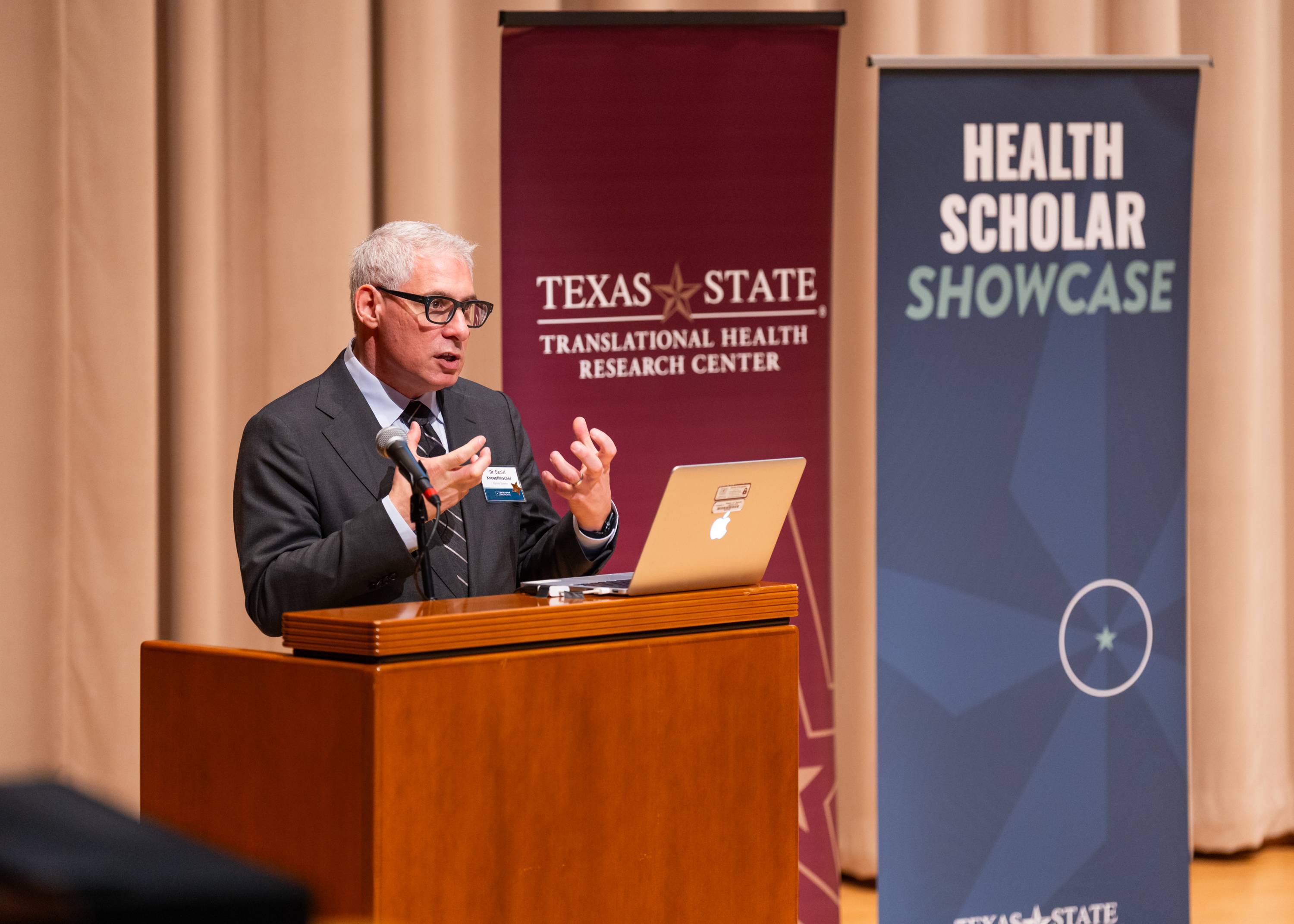 Dr. Daniel Knoepflmacher speaks at the podium. Behind him are banners for the Translational Health Research Center and Health Scholar Showcase