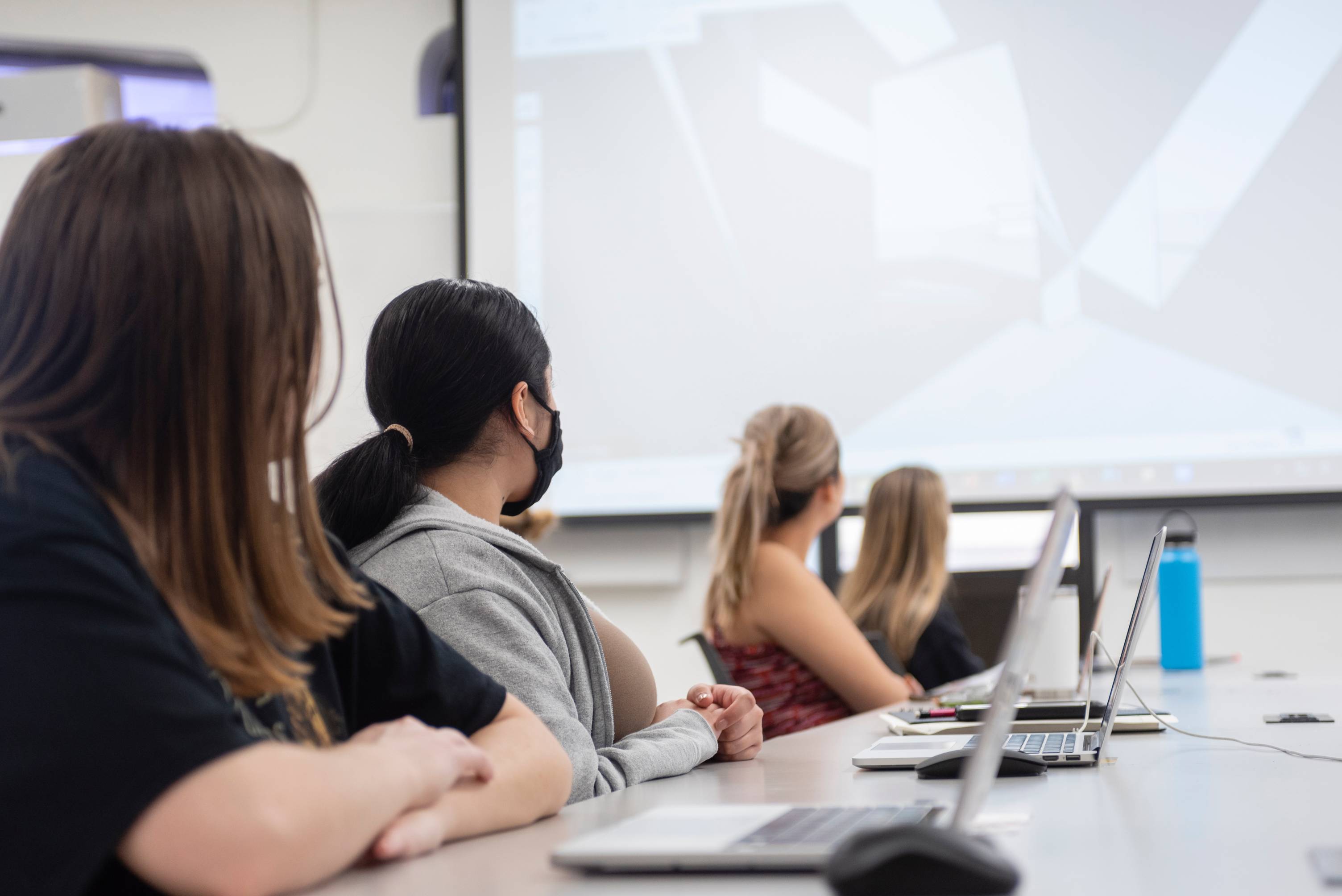 Students sit at a table with laptops out in front of them. The students are looking towards a screen projection at the front of the classroom.