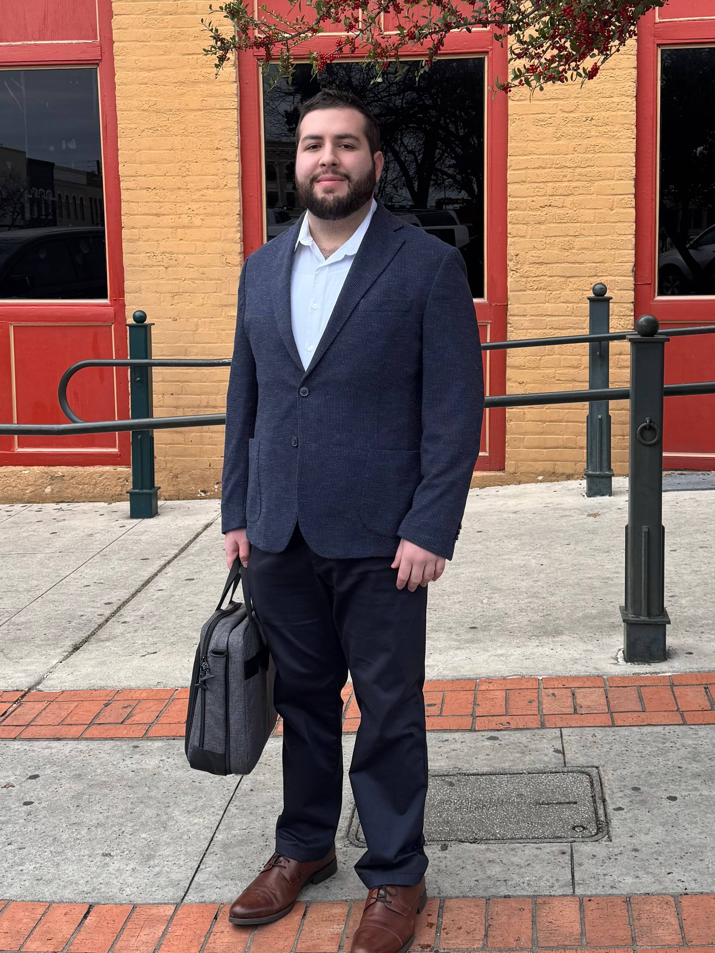 Zachary is standing on the sidewalk. He is wearing a professional blue coat, slacks, and black shoes. He is holding a grey bag in his right hand.
