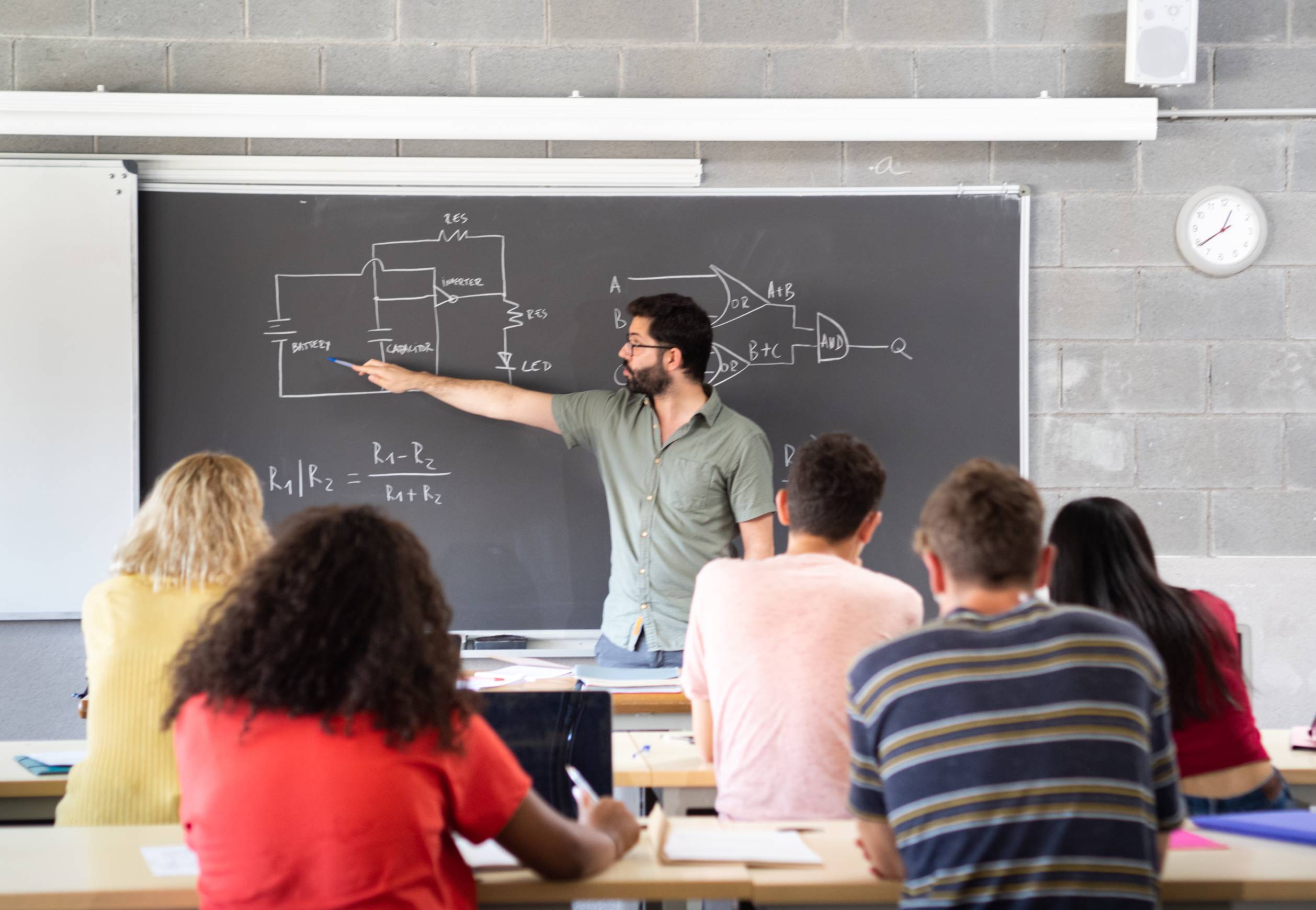Teacher pointing to a blackboard while students sitting on a desk.