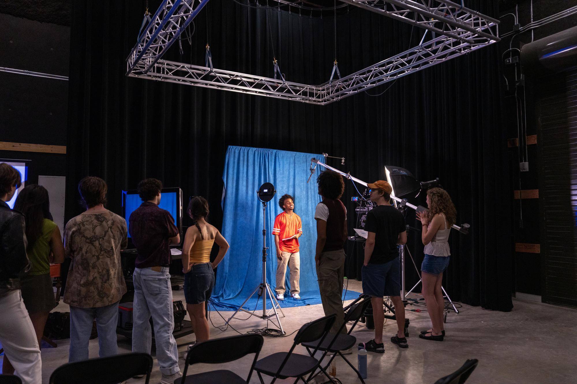 an actor in an orange shirt stands in front of a blue backdrop while film crew film him