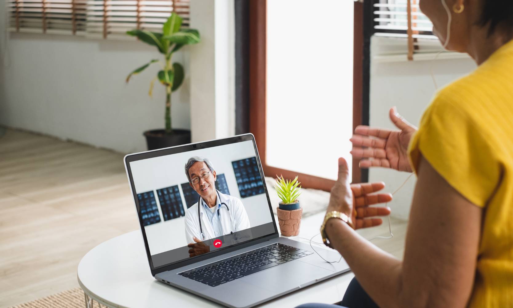 A woman an yellow is having a telehealth appointment with a doctor on her laptop.