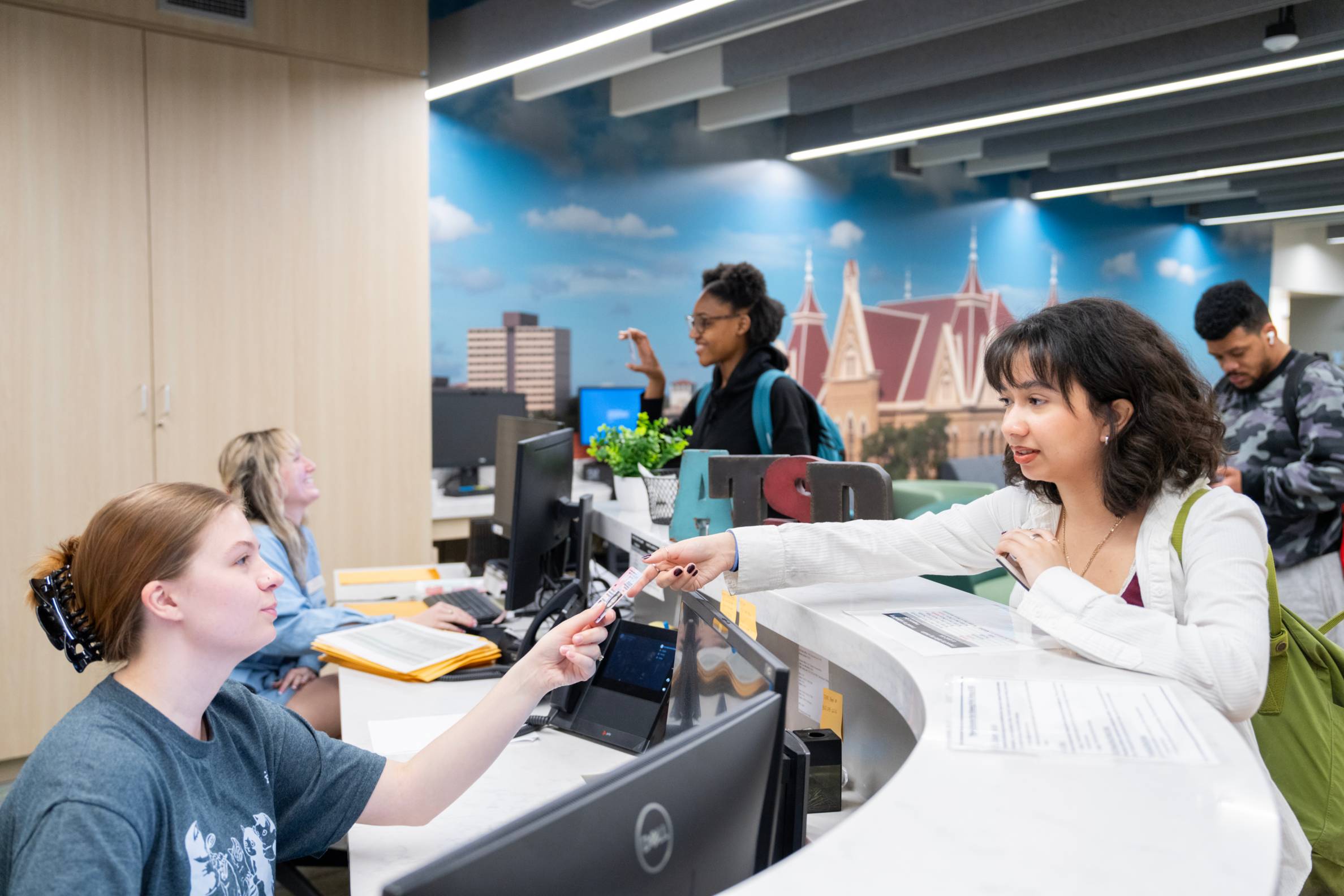 A student is reaching over a service counter desk providing the assistant with a card. On the service counter, decorative block letters "ATSD" are displayed.