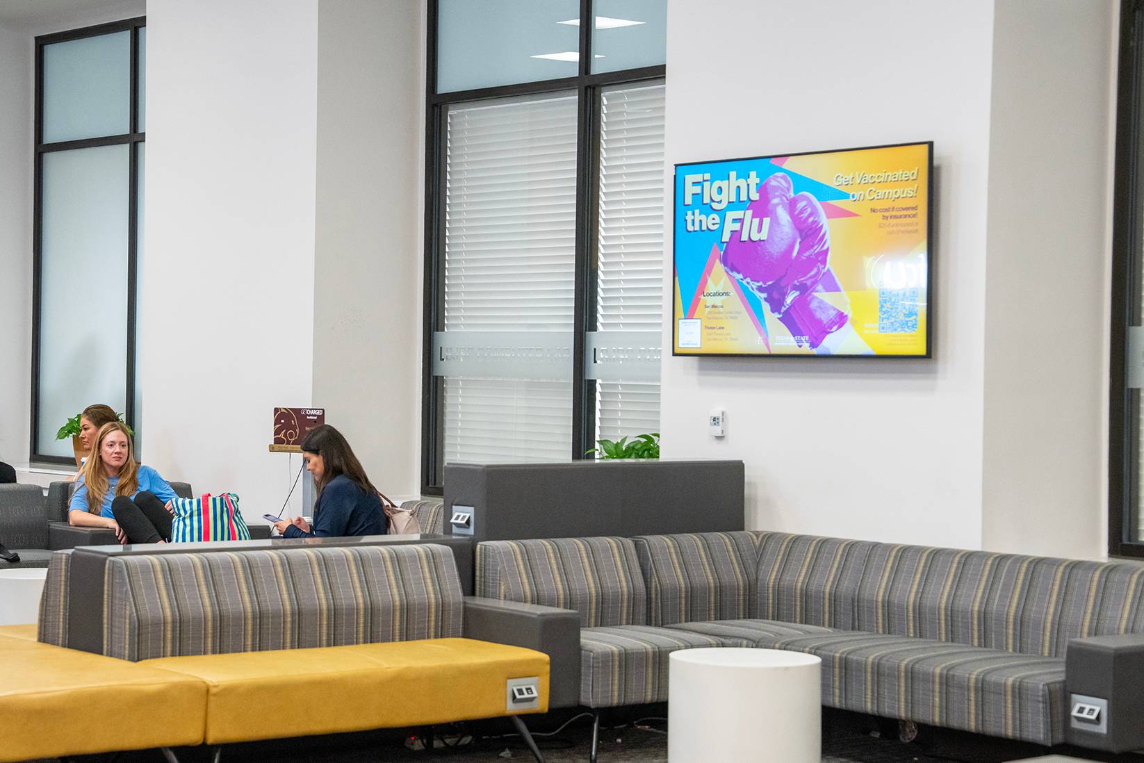 digital sign on wall above couches with students sittingon them