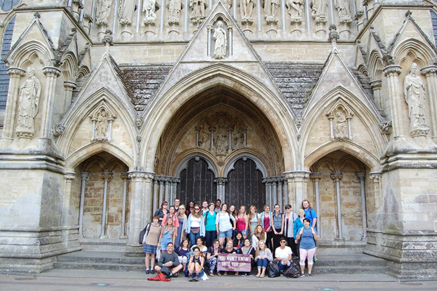 Texas State students group photo in front of ornate church in Canterbury, England. Group is holding a banner that says "You won't know until you go."
