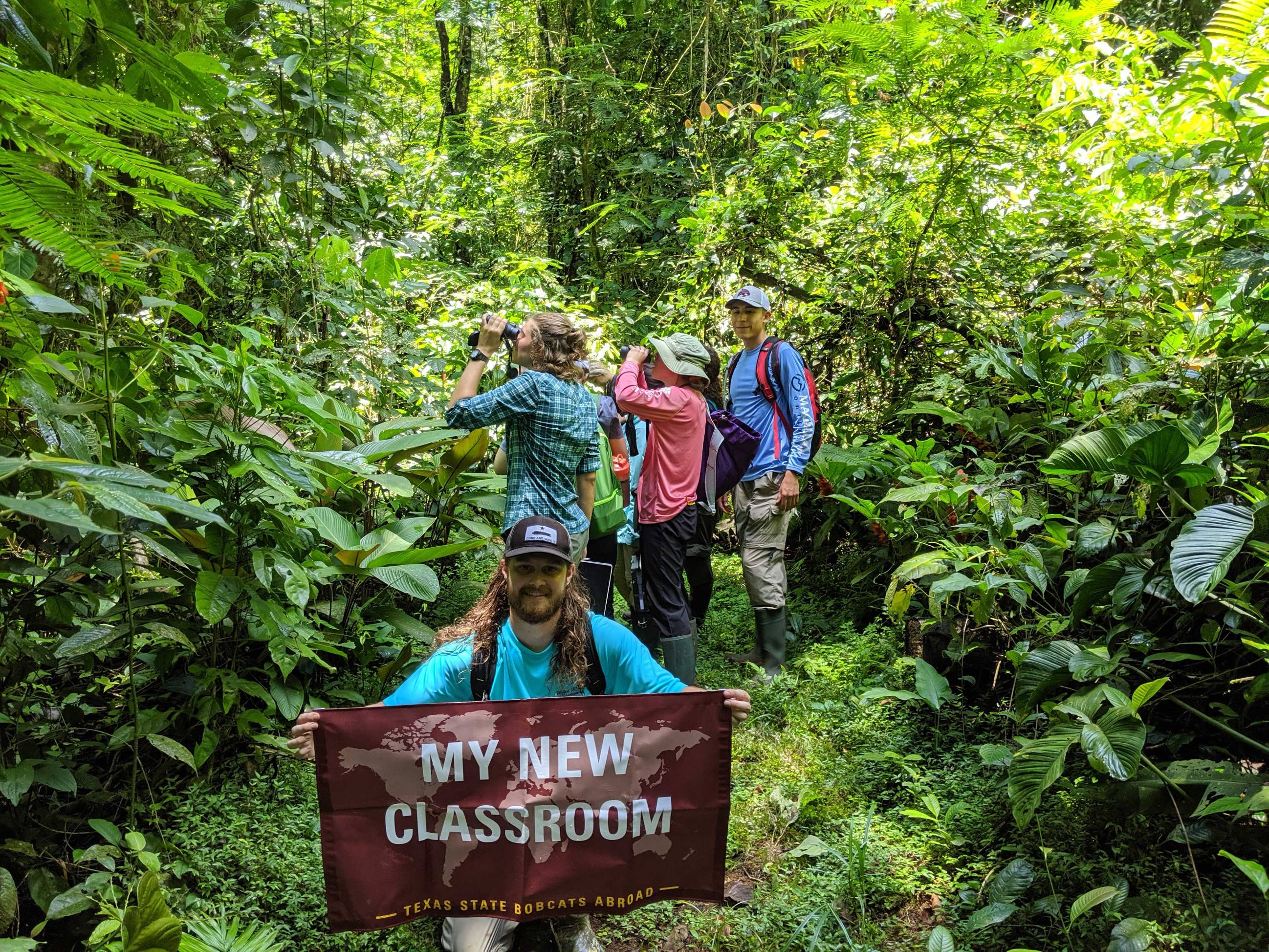 Texas State students in a forest in Costa Rica. Student is holding a banner from TXST Education Abroad that says "My new classroom."