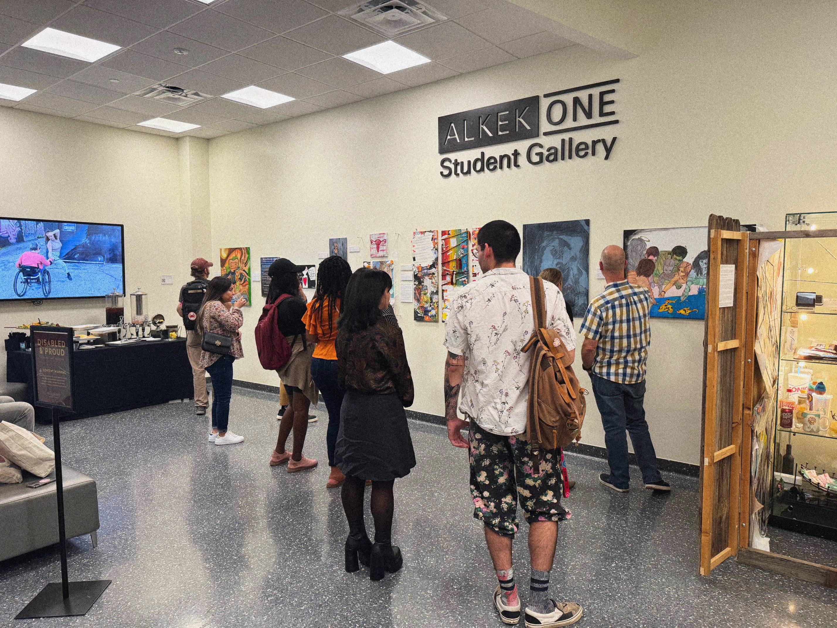 A photograph of the Disabled & Proud 2025 opening reception is displayed. Observers are in the space looking at art pieces which hang on the wall. Above the art, a sign reads "Alkek One Student Gallery". To the left of the crowd is a screen showing a video dance. A dancer utilizing a mobility aid is shown.