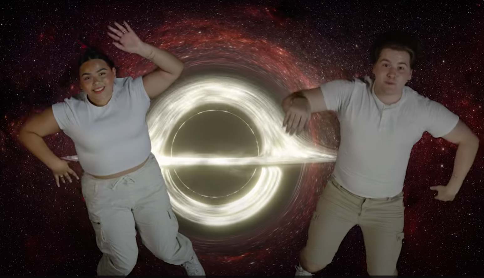 Two people in white shirts and light pants pose in front of a glowing black hole and star-filled space background