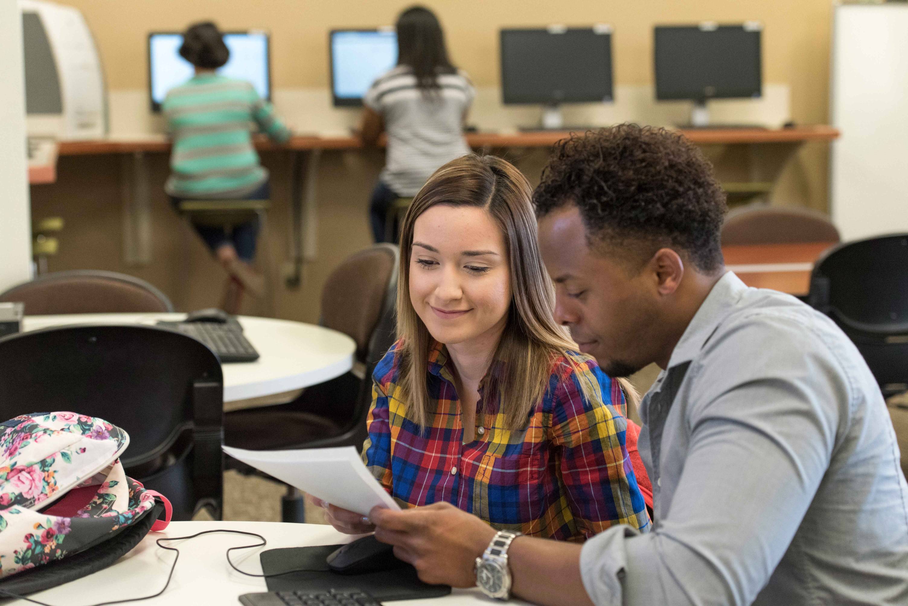 Two people sit together at a table in a computer lab, looking at papers, with computers and other people working in the background