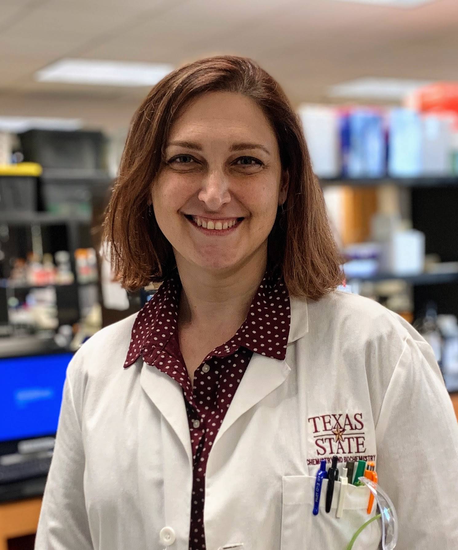 A faculty wearing a Texas State University lab coat stands in a laboratory, smiling at the camera. She has shoulder-length brown hair and is dressed in a maroon shirt with white polka dots underneath her lab coat.