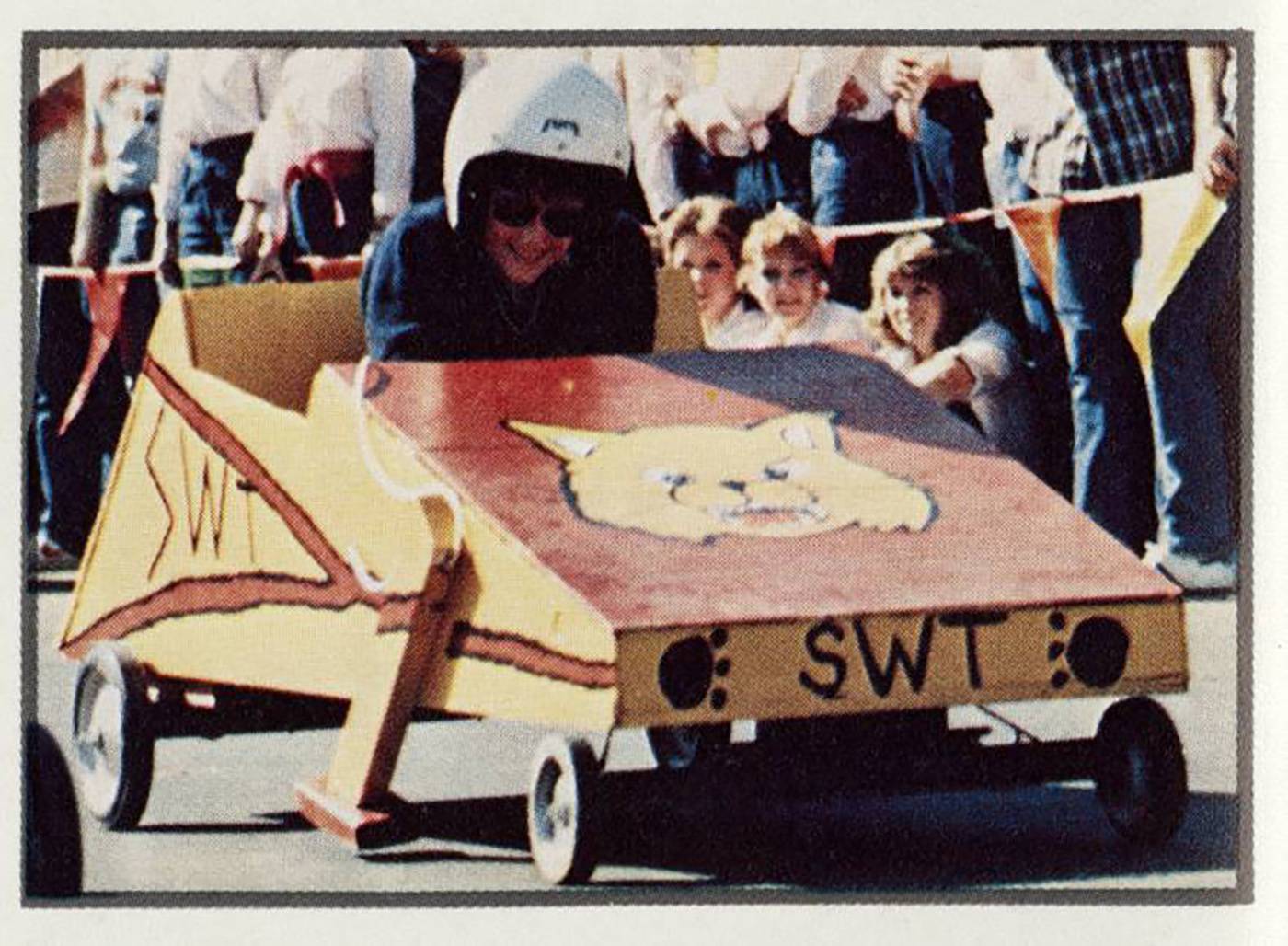 side profile of man in soap box derby car with bobcat painted on hood