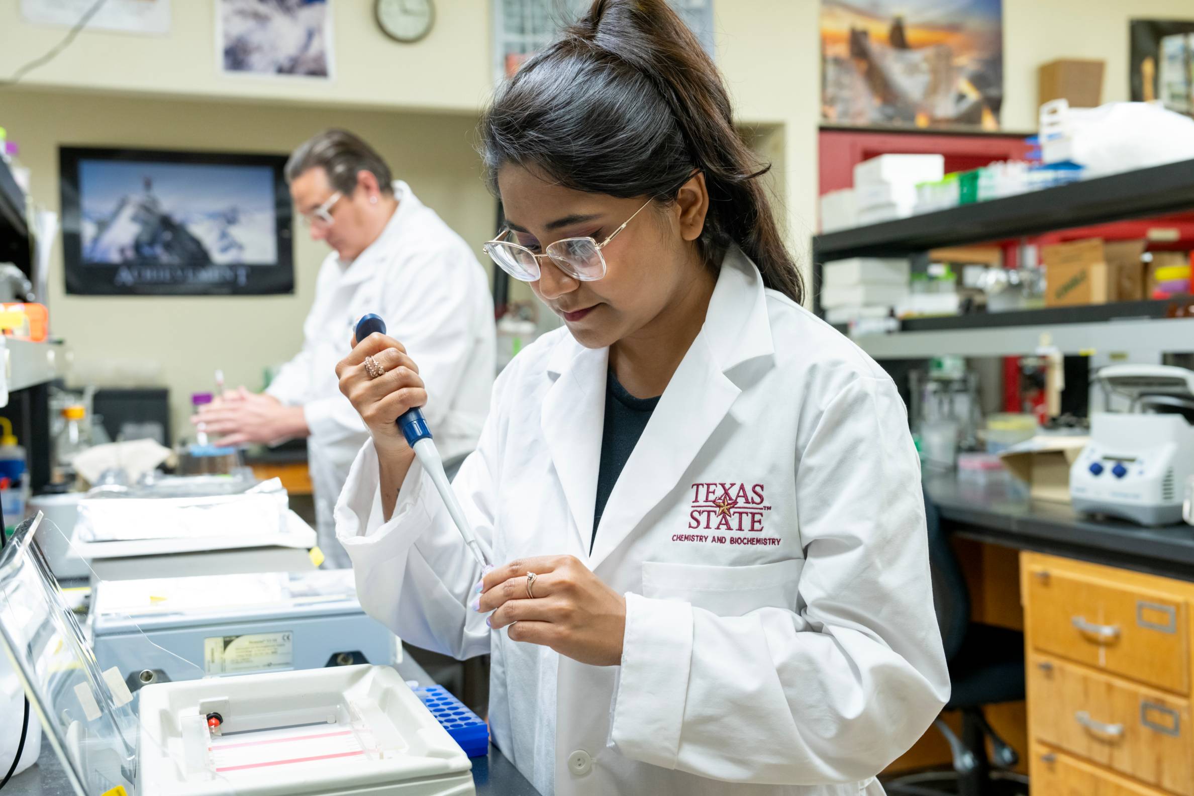 A student with her hair tied back and wearing glasses, a white lab coat with a "TEXAS STATE CHEMISTRY AND BIOCHEMISTRY" logo is intently using a pipette in a lab. Another person in a lab coat is visible in the background, out of focus.