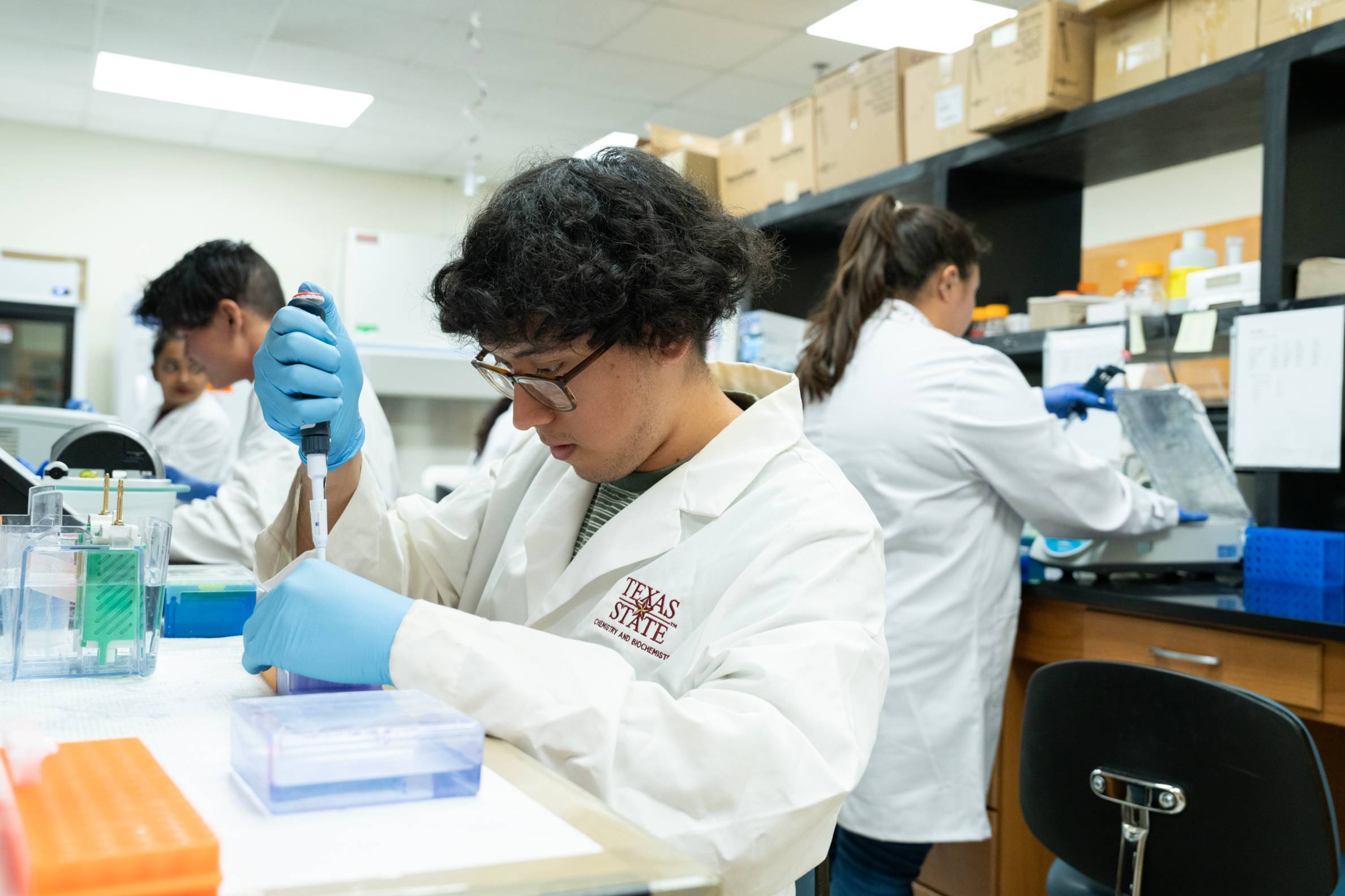 A student with curly hair, wearing a white lab coat with "TEXAS STATE" on it, blue gloves, and safety glasses, meticulously dispenses liquid with a pipette into a small tube. In the background, other students in lab coats are also working.