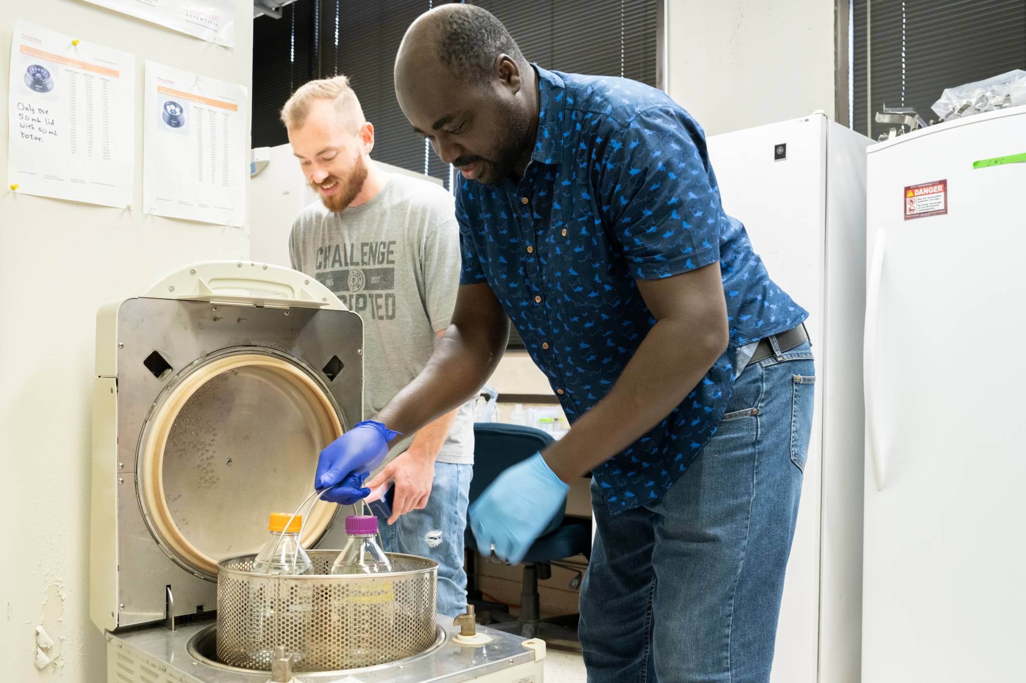 Two researchers work together in a laboratory. One, wearing a blue patterned shirt and gloves, places flasks with orange and purple caps into a centrifuge.