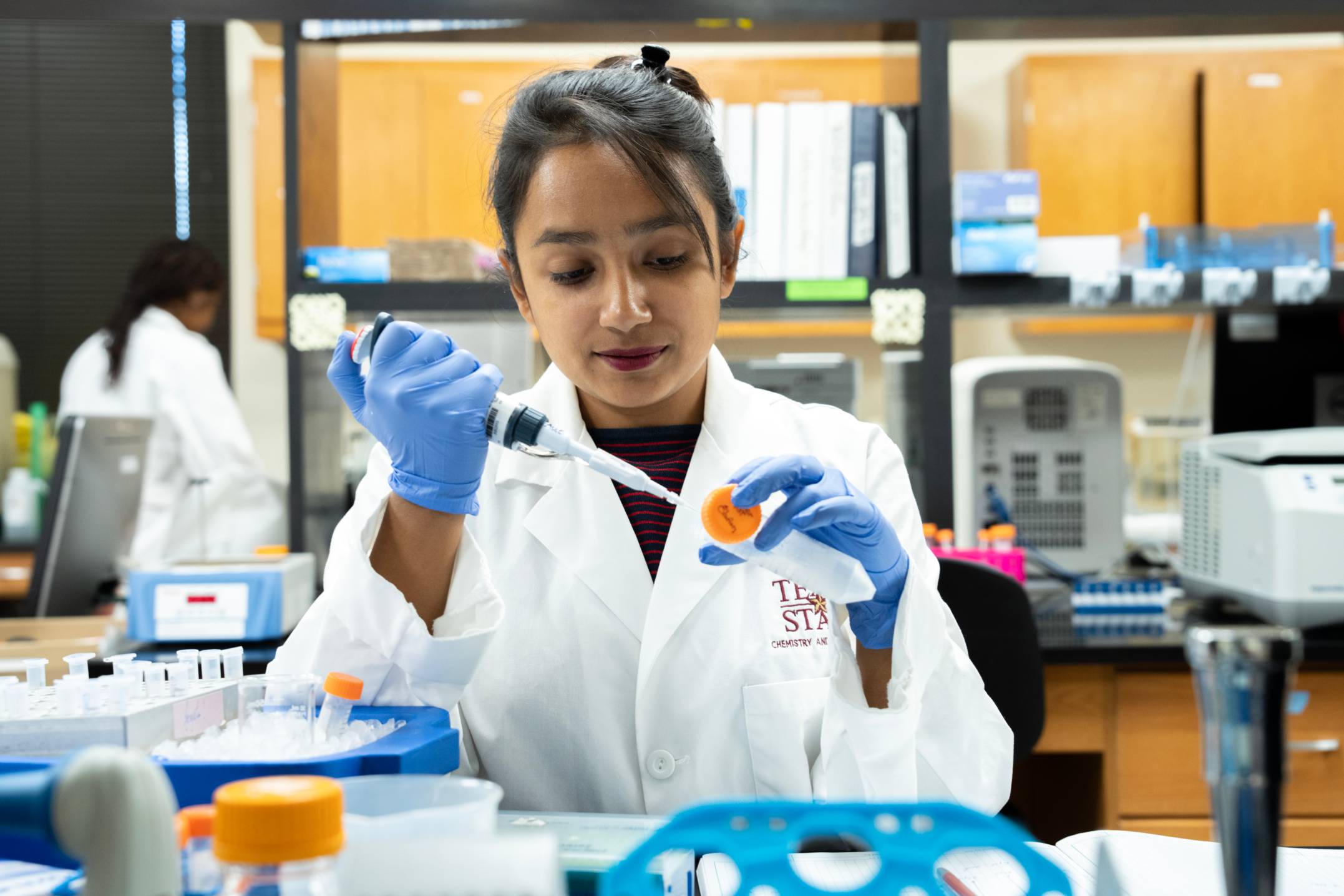 A student in a white lab coat and blue gloves uses a micropipette to transfer liquid into a small tube in a laboratory. 
