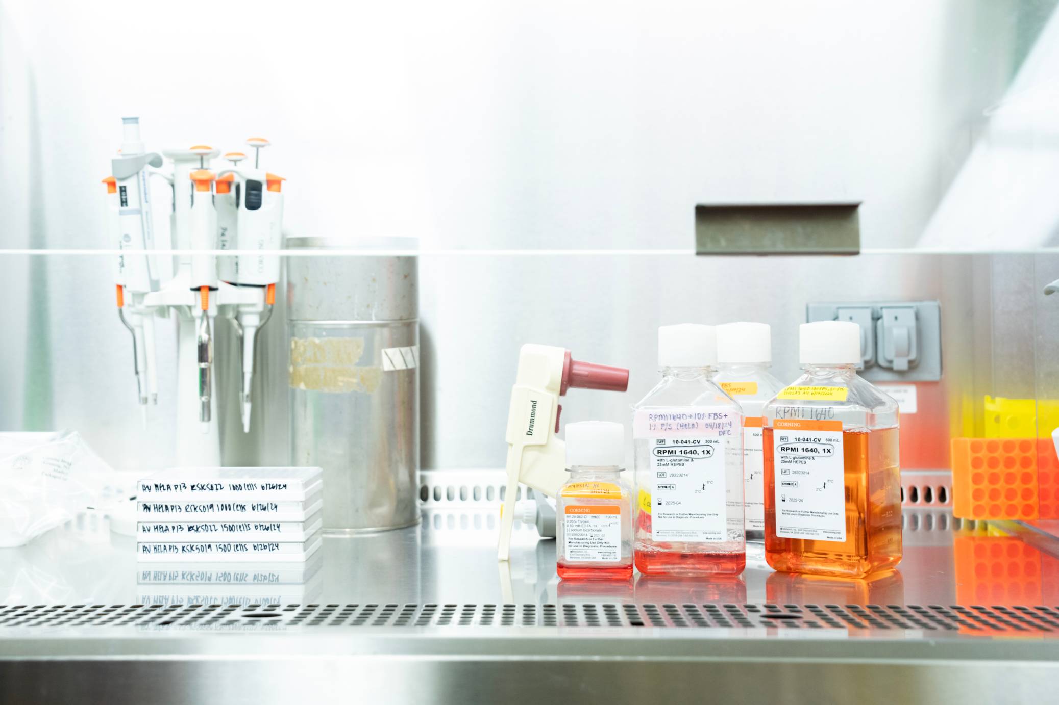 A lab bench is shown from a low angle, featuring various scientific instruments and bottles. On the left, several pipettes are arranged in a stand, and a stack of white petri dishes sits on a white rectangular base. In the center, a metal container stands next to a small, white, and orange bottle labeled "Brand". To the right, three clear bottles with orange or red liquids are labeled with white stickers, sitting in front of yellow-orange containers holding multiple test tubes. The background is a blurry white wall.