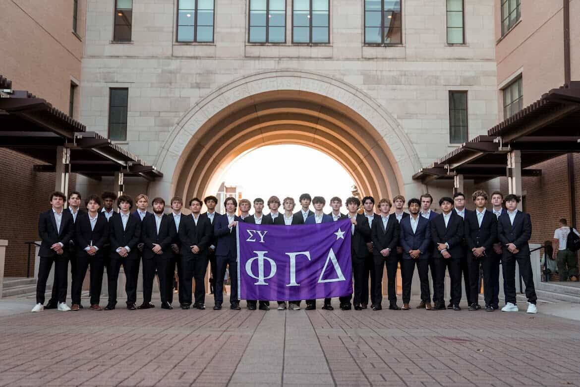 Men of Phi Gamma Delta (FIJI) stand behind a table and under a canopy while recruiting in the Quad