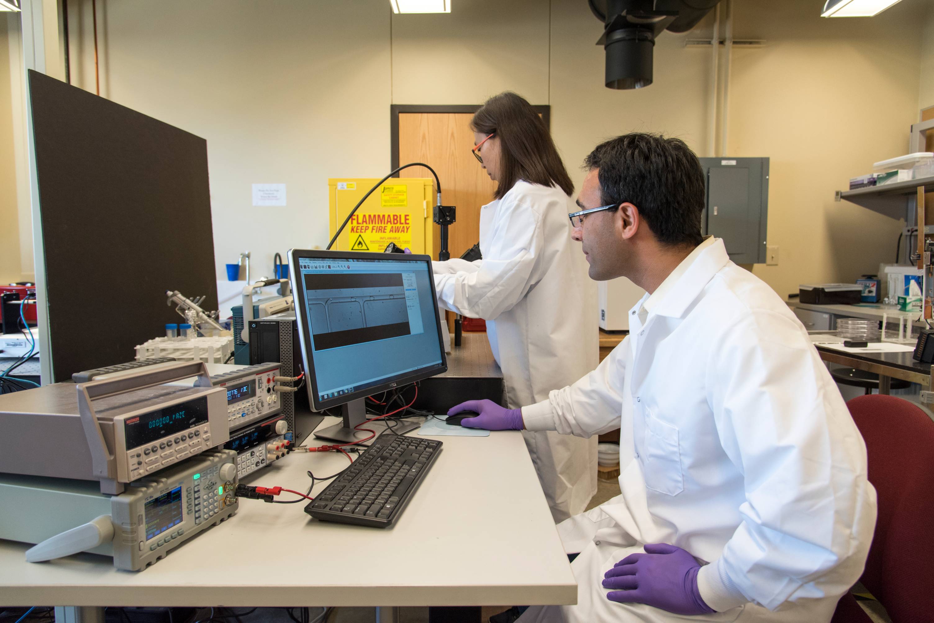  Two researchers in lab coats work in a laboratory equipped with advanced instrumentation. One person operates a computer displaying microscope images, while the other adjusts a scientific apparatus connected to the system.