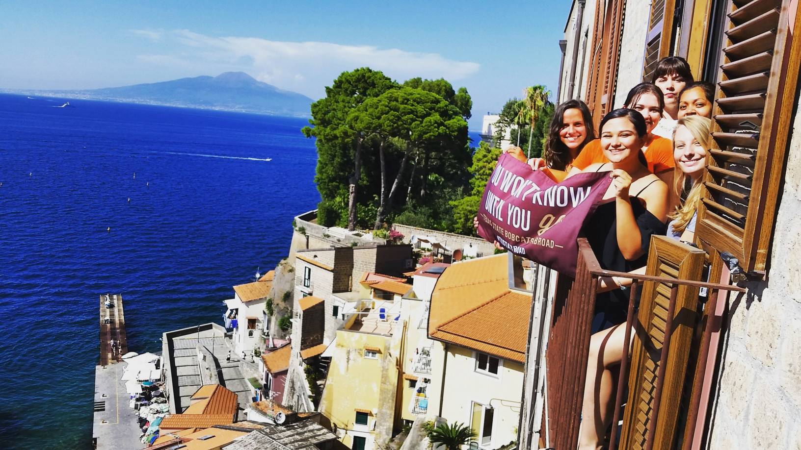 Students holding an education abroad flag above a balcony overlooking the mediterranean sea.
