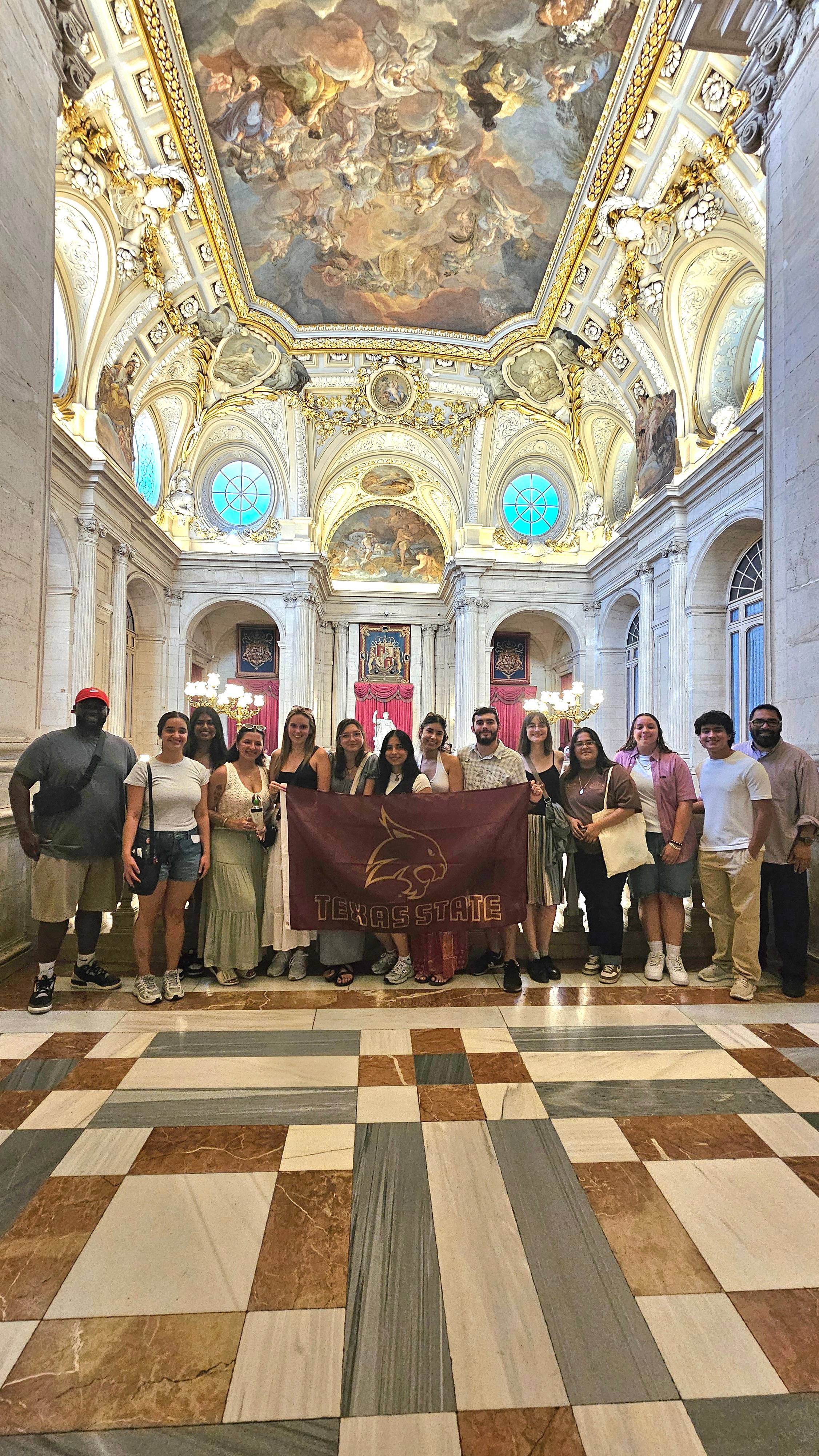 Group of students standing inside the Madrid Royal Palace with a TXST flag.