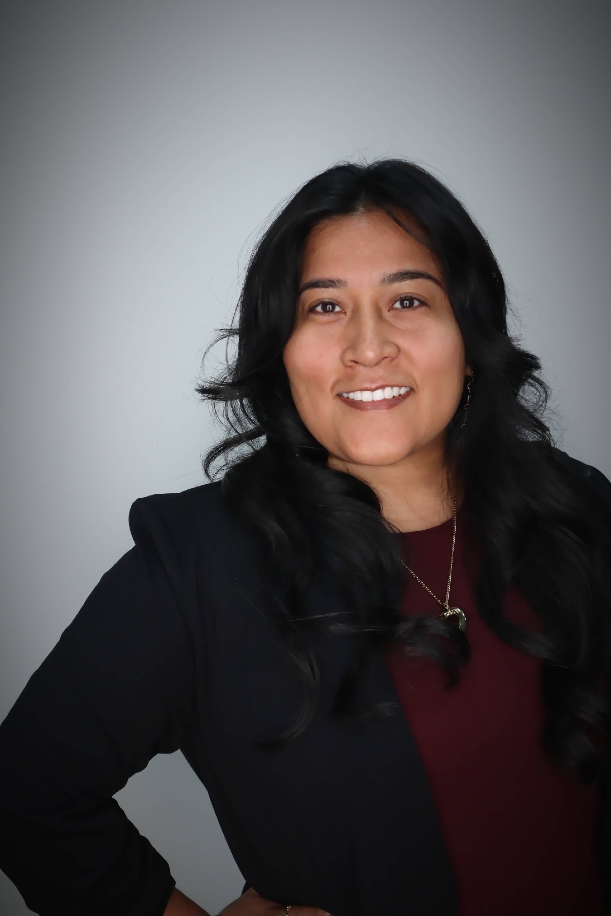 Maria De Los Santos Headshot. Maria smiling, wearing a black blazer over a maroon shirt.