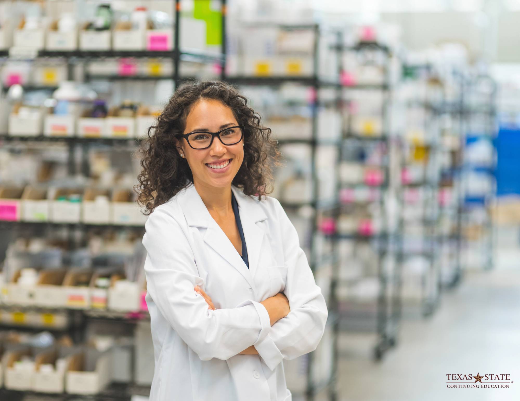 Pharmacist standing, cross-armed, in front of rows of prescription medicine
