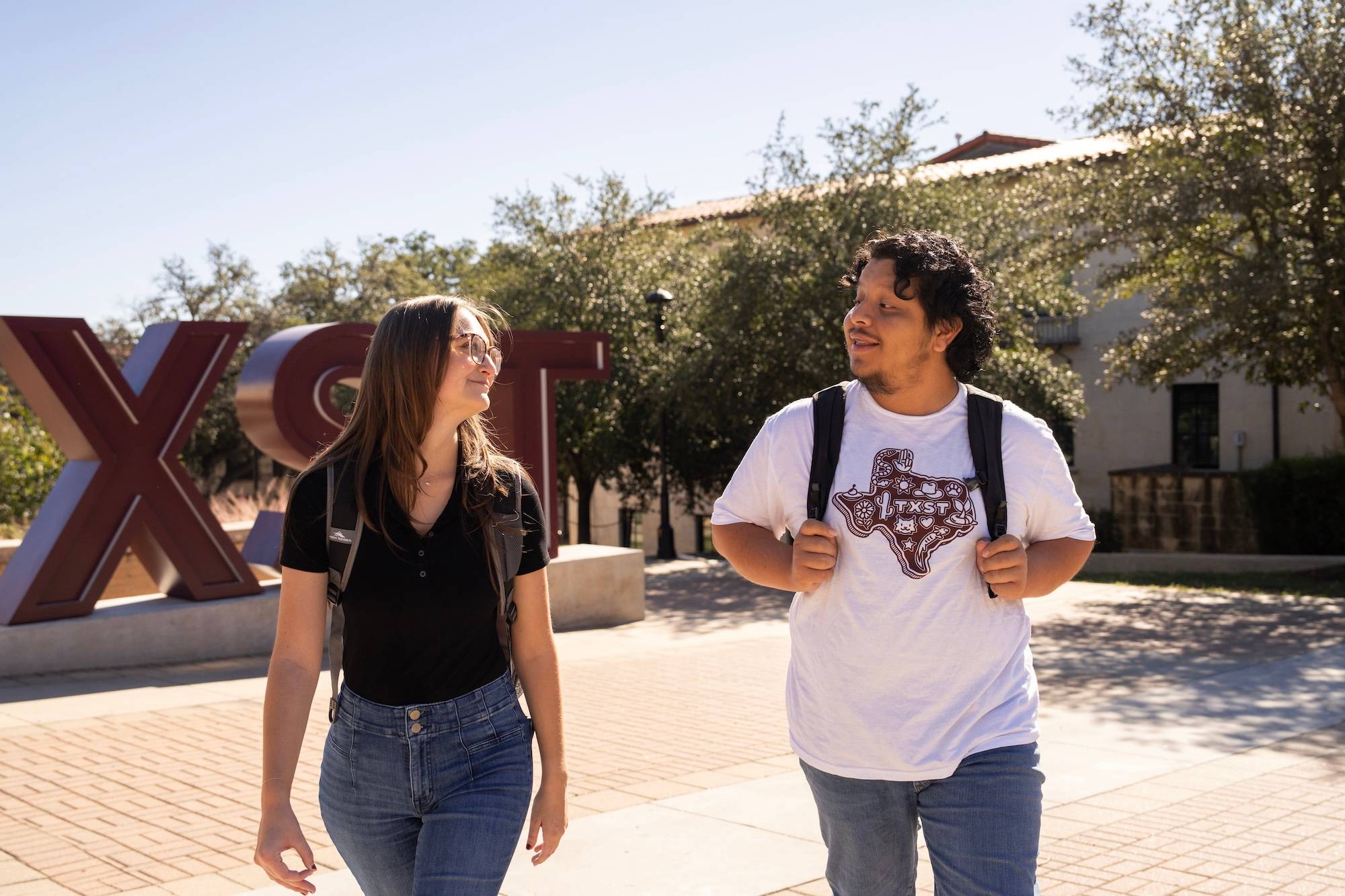 Two students talking and walking on campus next to the TXST sign