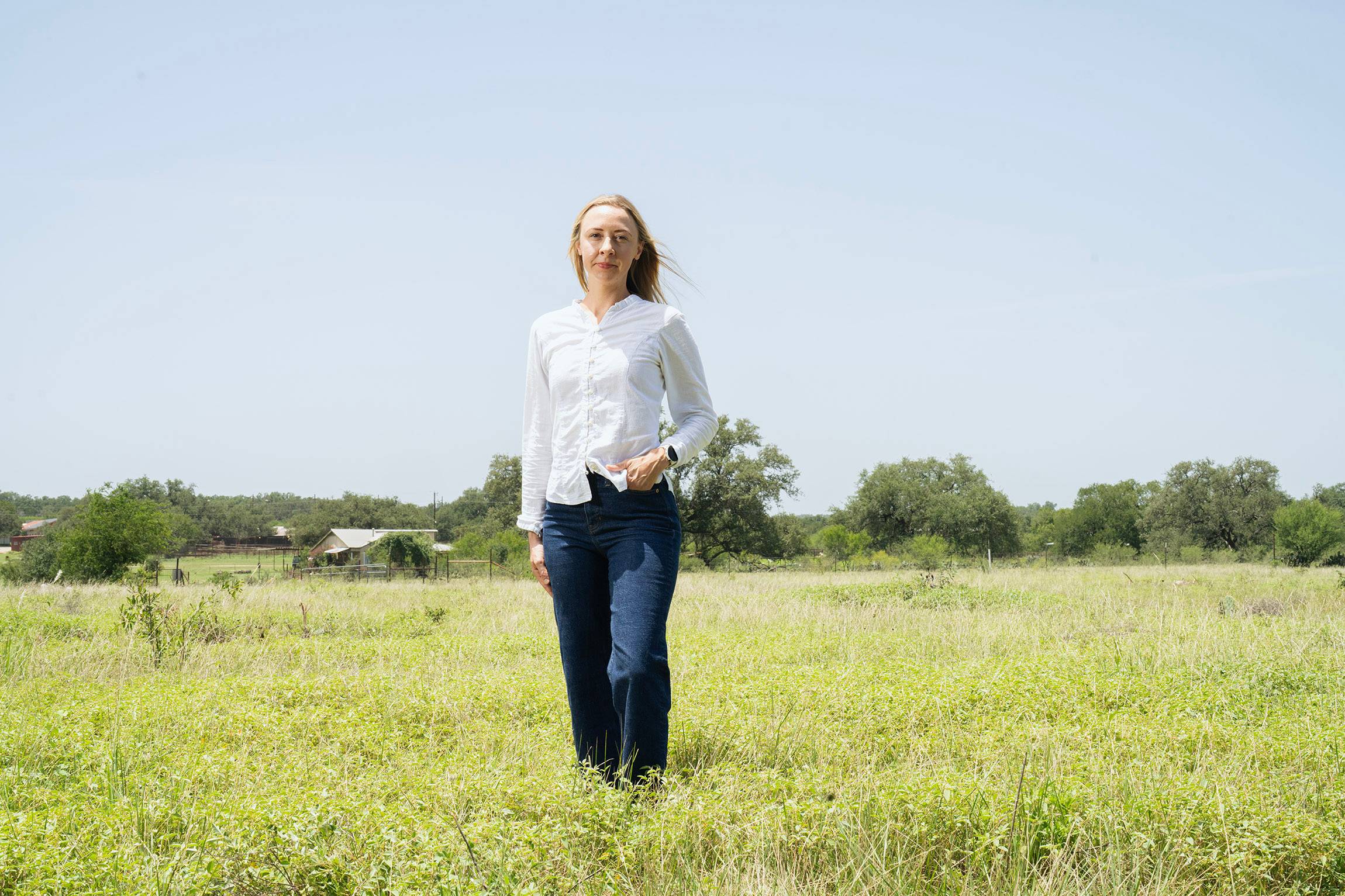 Woman dressed in a white sleeved shirt and jeans standing in the middle of a green field with trees in the background.