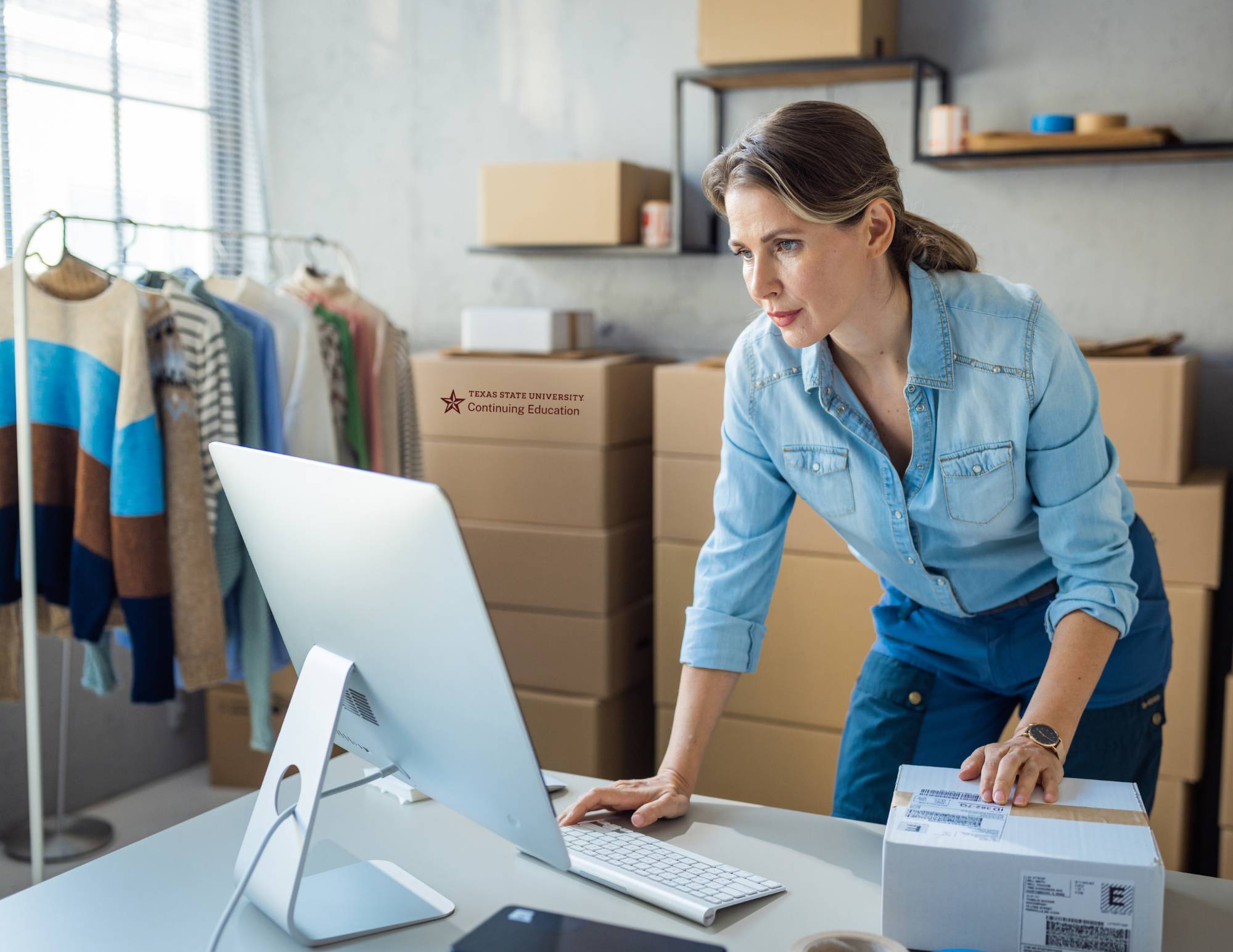 a woman in a small business typing on a mac while placing her hand on a shipping box