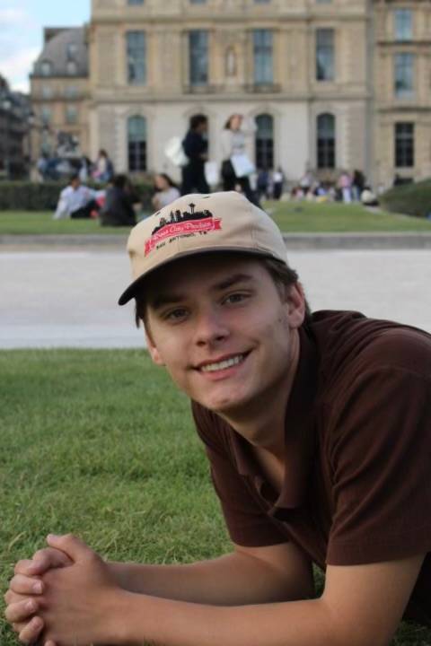 man lying on grass, propped on elbows, wearing a red shirt and a baseball cap, palace in background