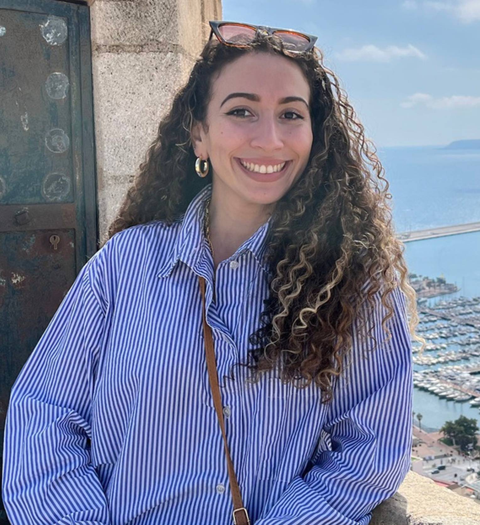 in striped shirt stands by stone structure labeled 'DE LA CAMPANA'; marina and sea in background.