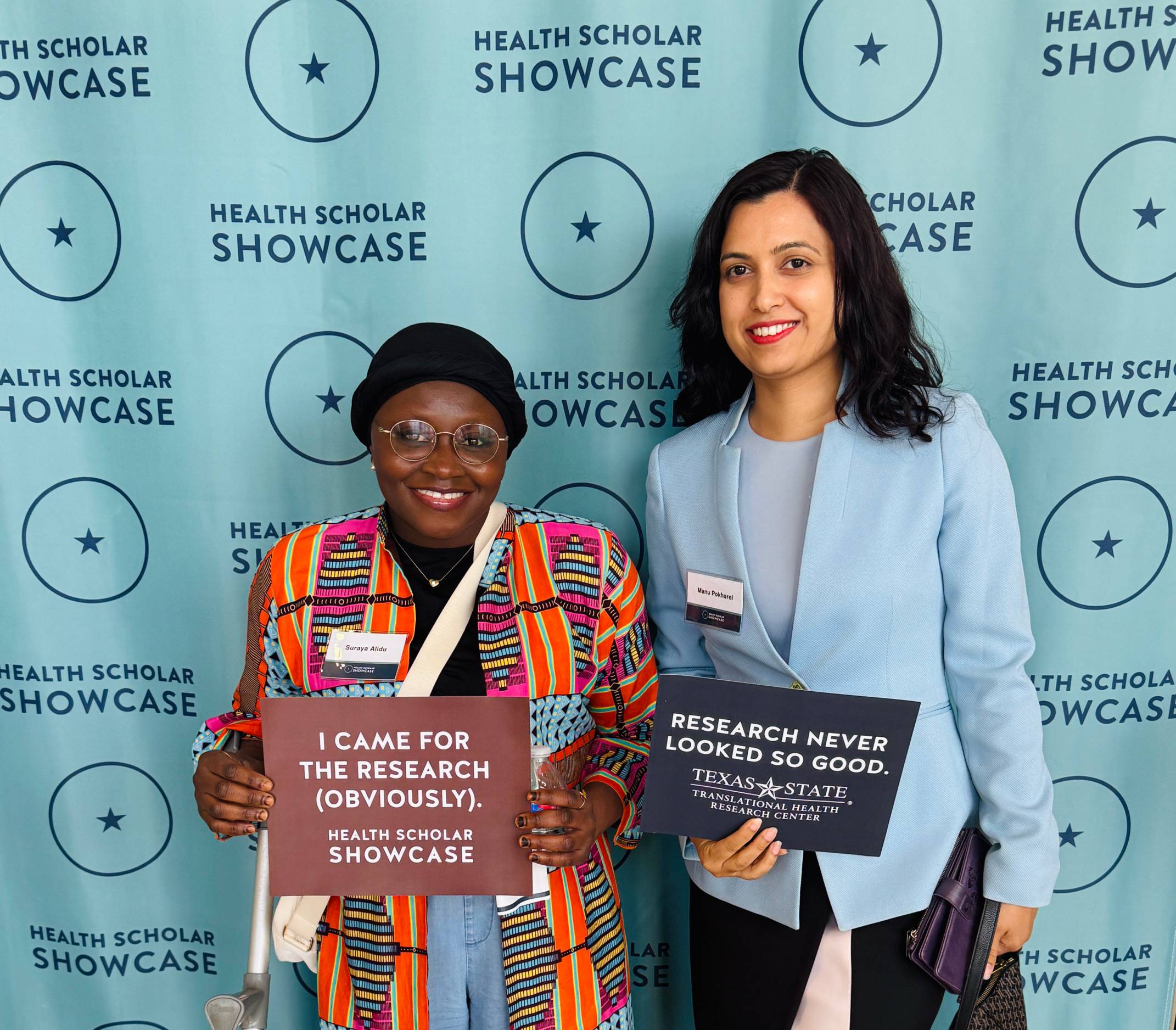 two women standing in front of large teal banner noth holding upsign for health scholar showcase