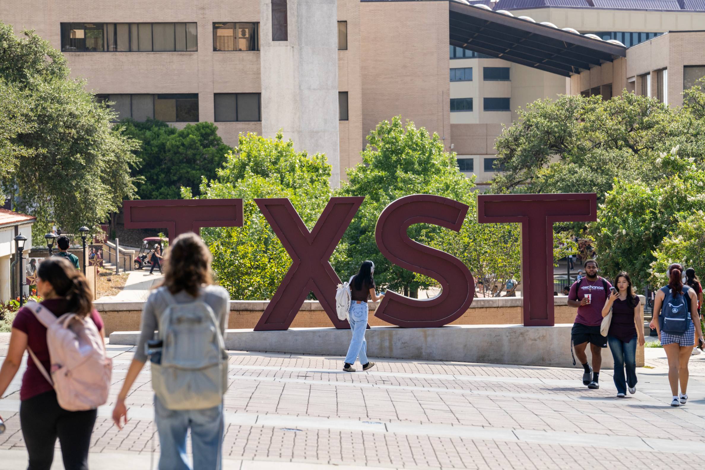 TXST sign in maroon next to students walking the campus