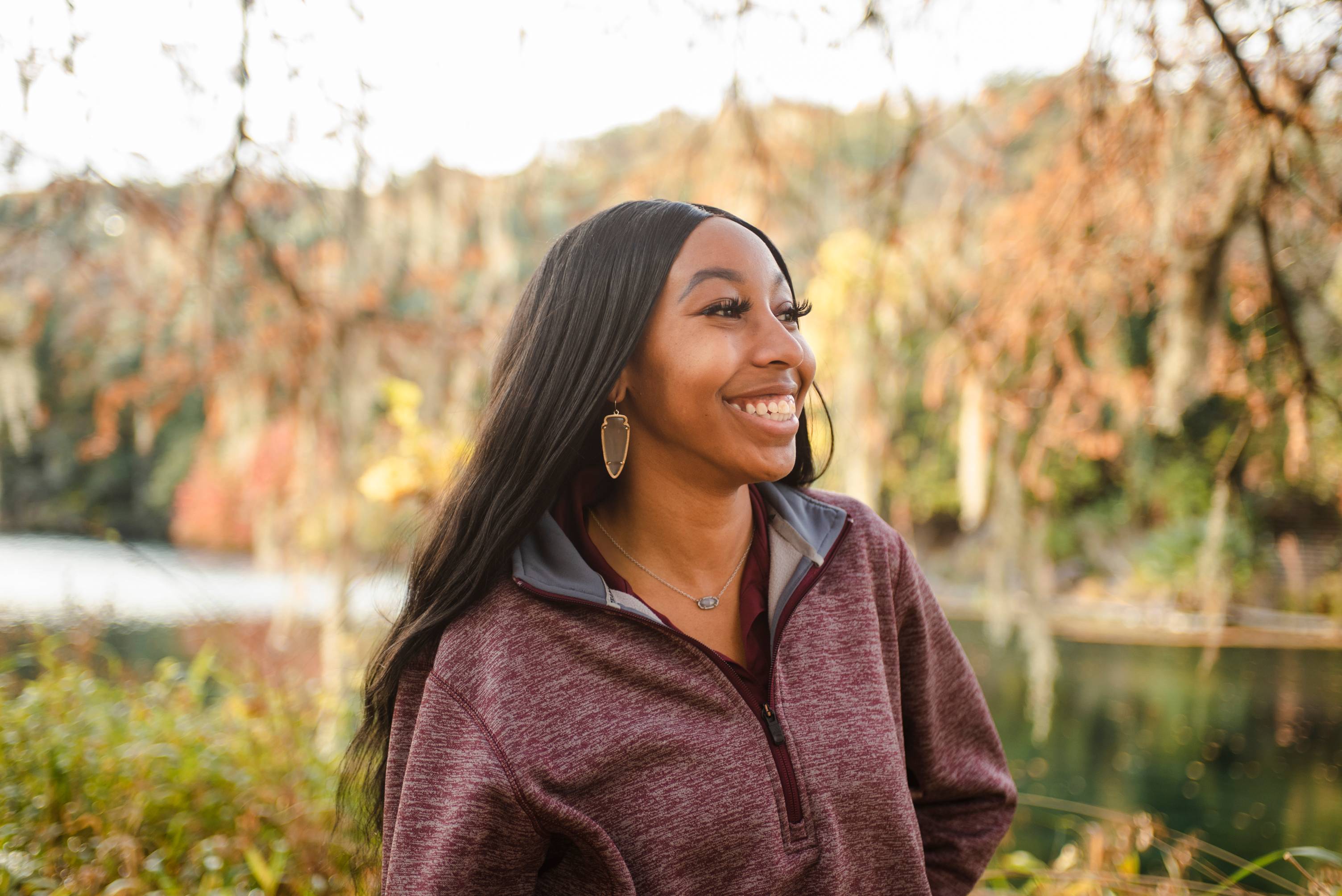 A black woman wearing a maroon TXST jacket and arrowhead earrings smiles into the distance with fall foliage in the background.