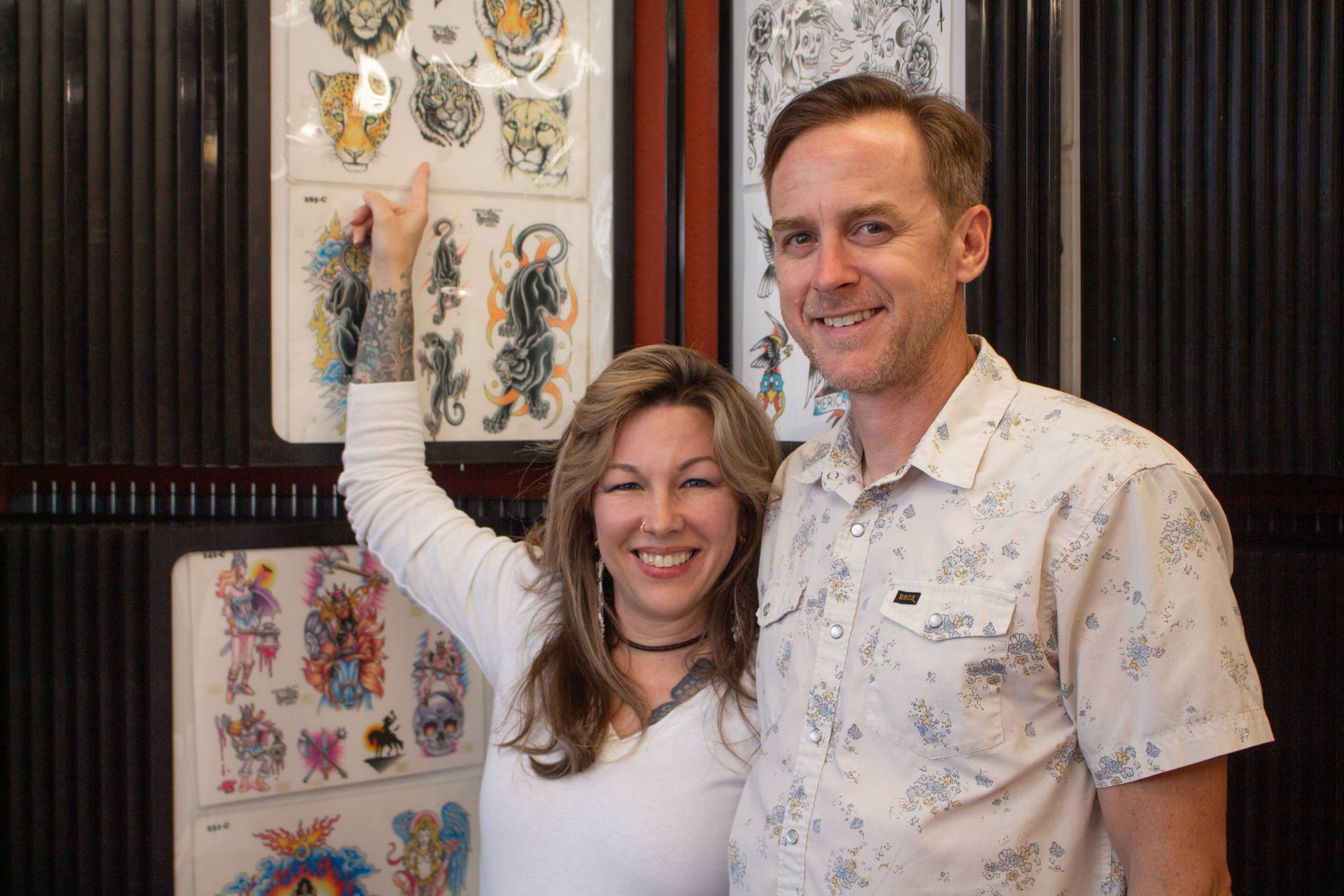 woman and man standing next to tattoo flash sheets on wall. woman is pointing to a bobcat tattoo