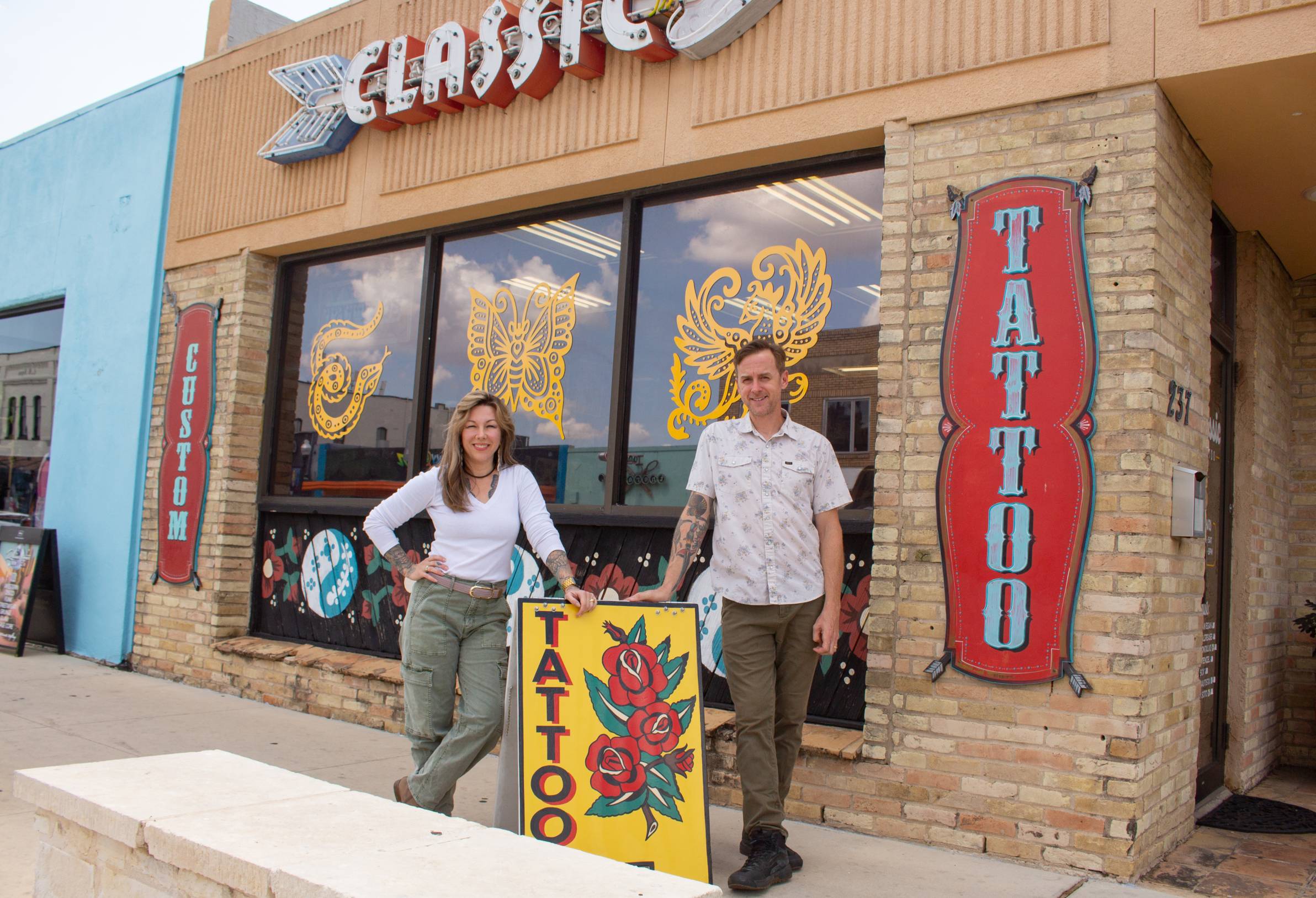 woman and man stand outside of a tattoo studio next to a painted yellow sign