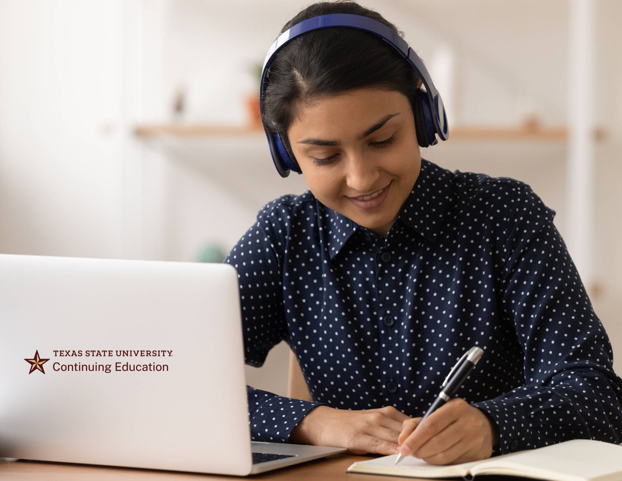 woman on headphones writing on paper next to laptop